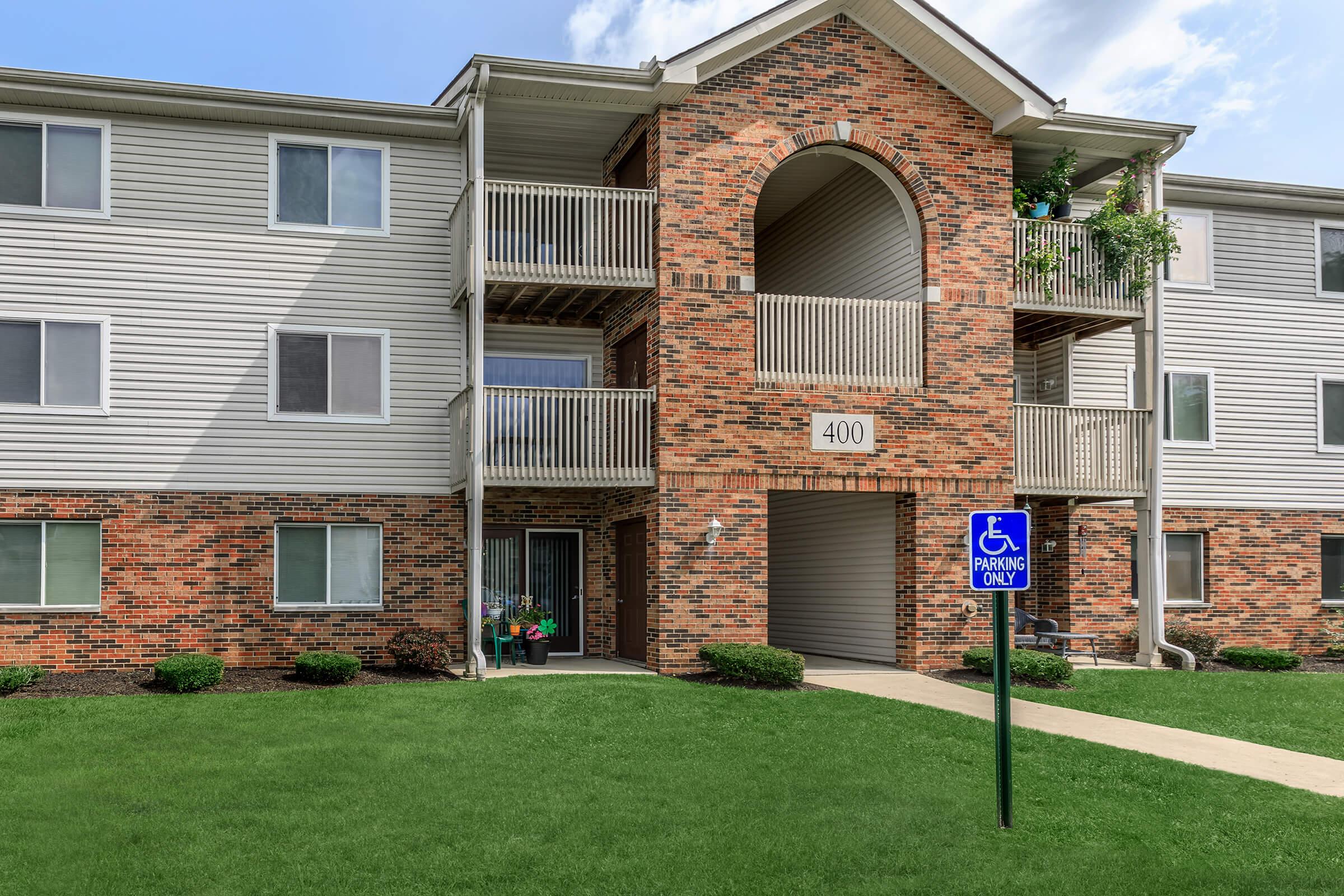 A brick and siding apartment building featuring an entrance with a large arch, balconies above, and a green lawn in front. A sign indicating "Handicapped Parking Only" is visible in the foreground, alongside a paved walk leading to the entrance.