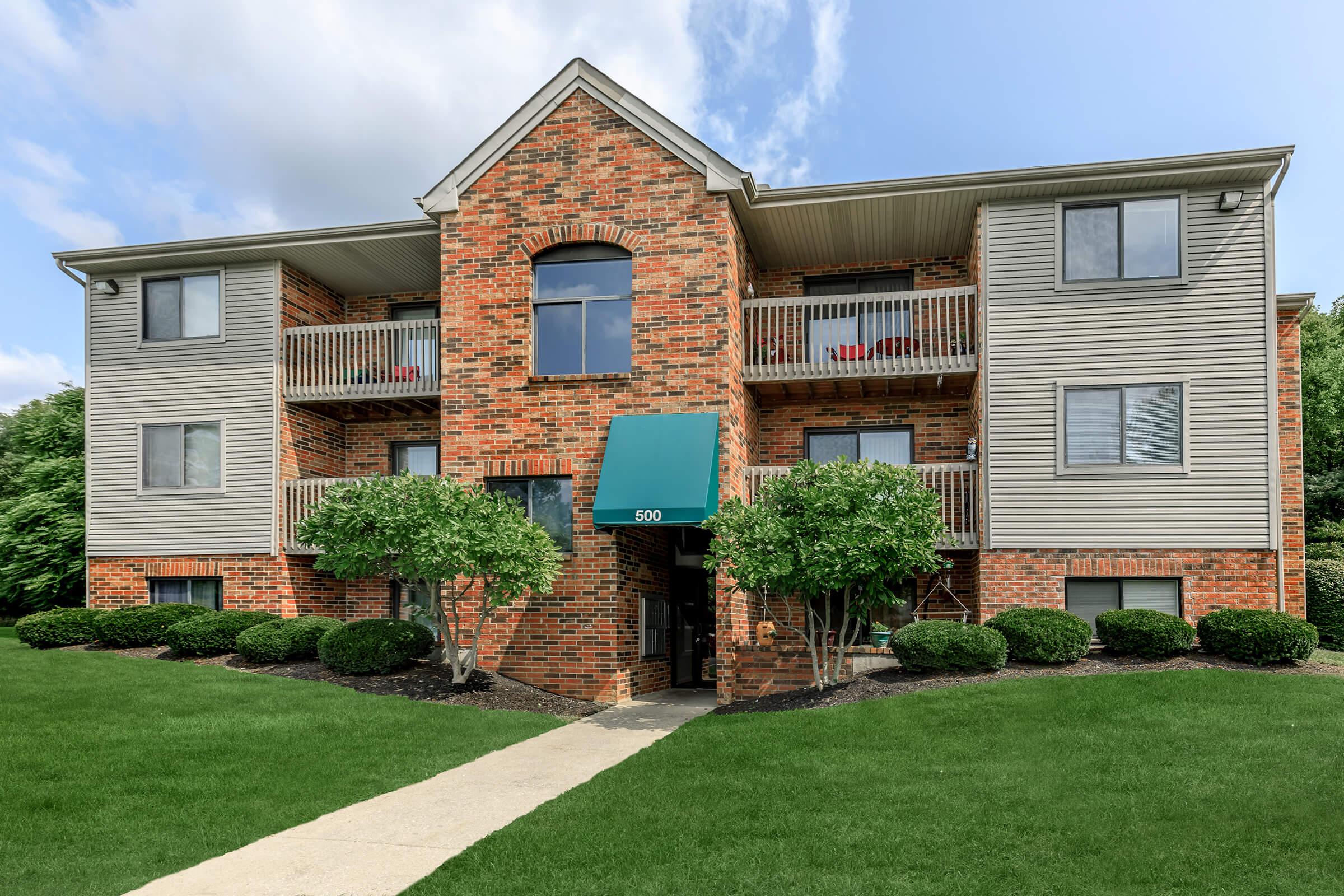 A three-story brick apartment building with gray siding, featuring a central entrance beneath a teal awning. Two small trees flank the entrance, and the lawn is well-maintained with a pathway leading to the door. Several balconies can be seen on the upper floors. Bright blue sky and fluffy clouds in the background.