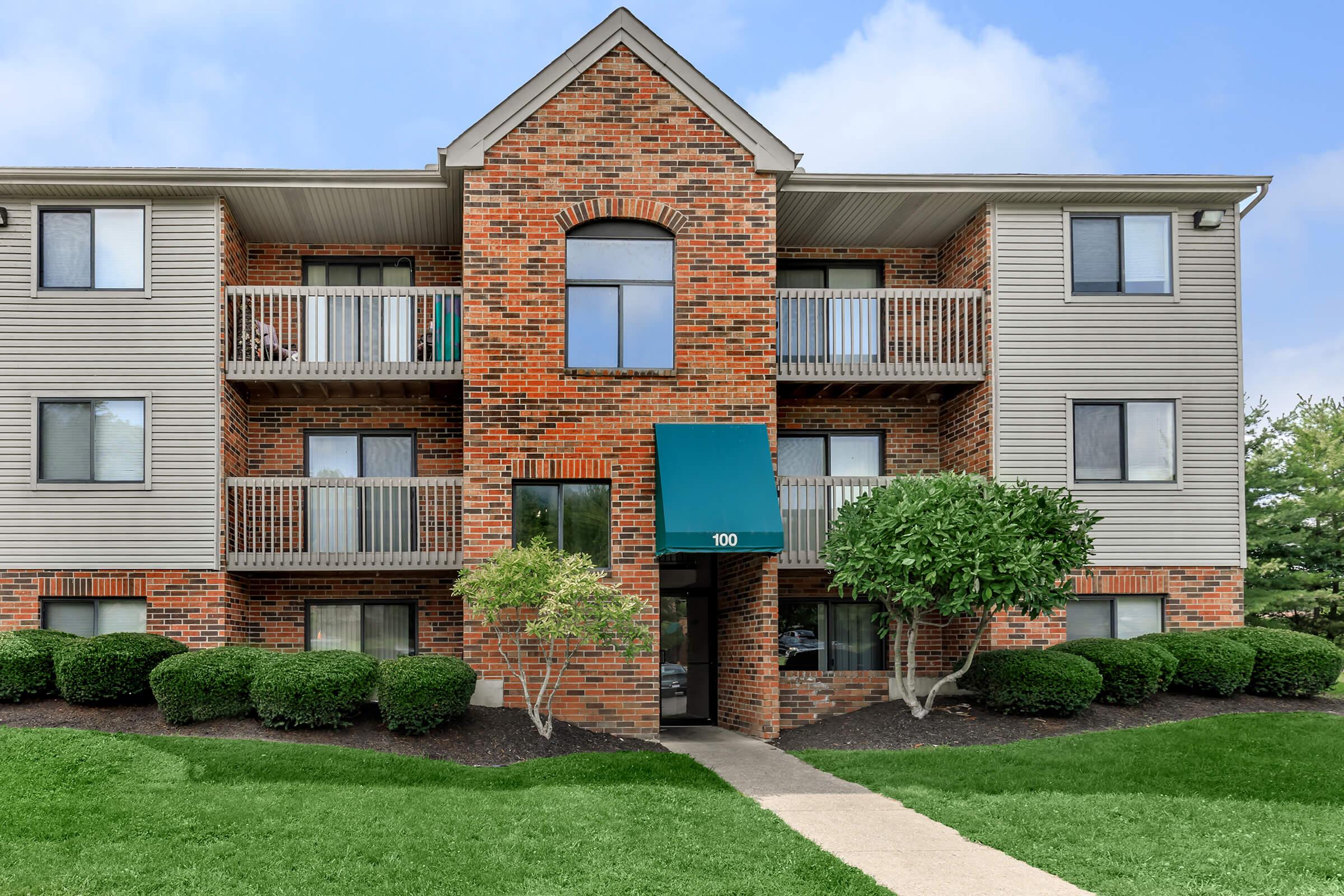 Brick apartment building with three stories, featuring a central entrance and a teal awning. The building has balconies on the second floor, surrounded by neatly trimmed bushes and lush green grass. The sky is clear with some clouds, creating a bright and inviting atmosphere.