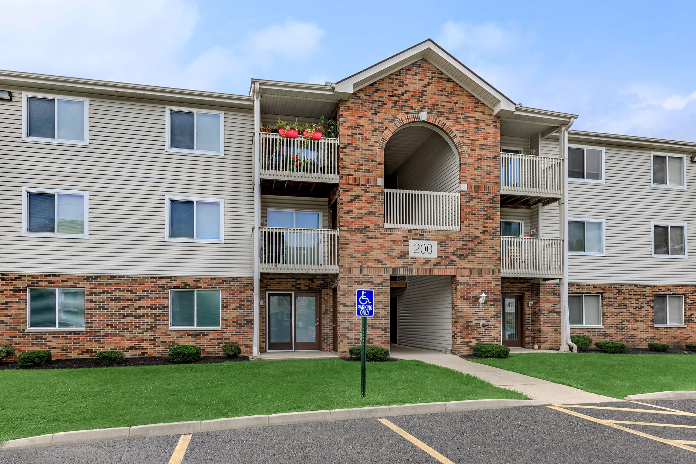 A three-story apartment building featuring a brick exterior and a modern design. The entrance is marked with the number "200" and includes a balcony with flower pots. There is a landscaped area in front and designated handicapped parking nearby. The sky is clear with minimal clouds.