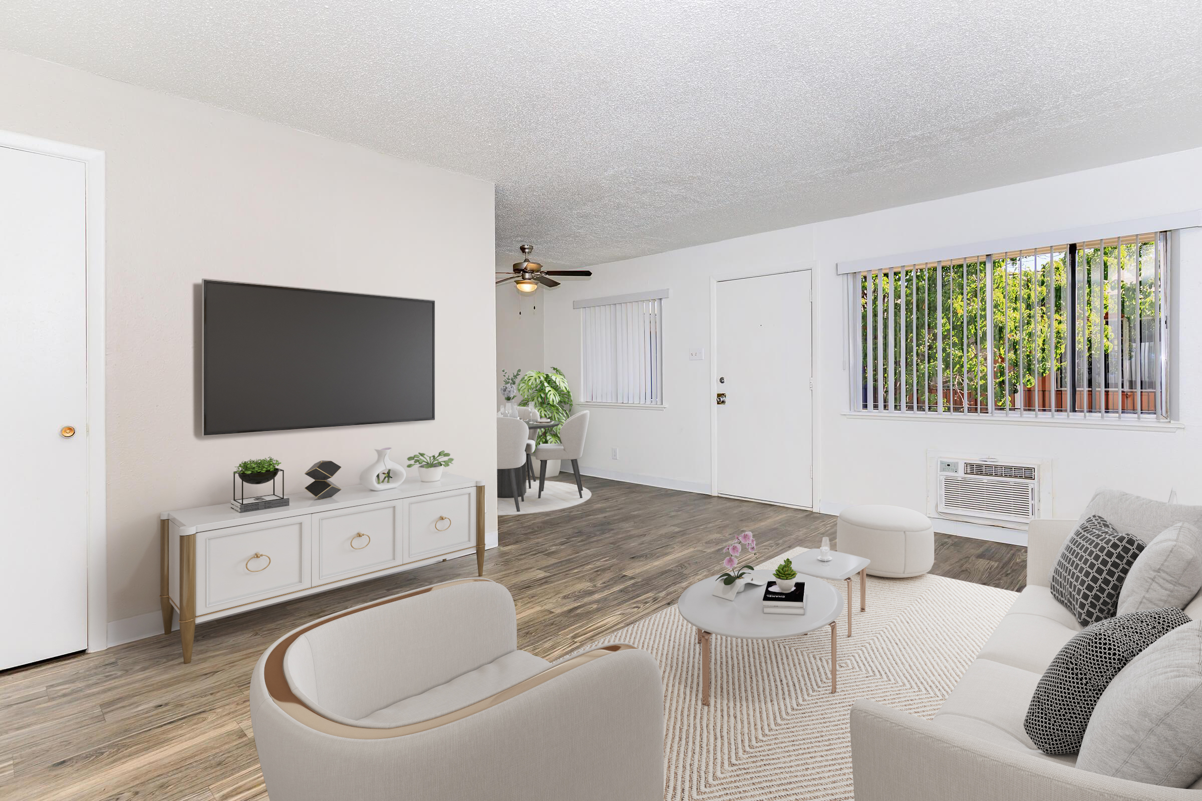 A cozy living room featuring a light-colored sofa and armchair around a round coffee table. A modern TV mounted on the wall above a white cabinet. A ceiling fan and bright natural light from a window with blinds, enhancing the open feel. An air conditioning unit is visible, along with a dining area in the background.
