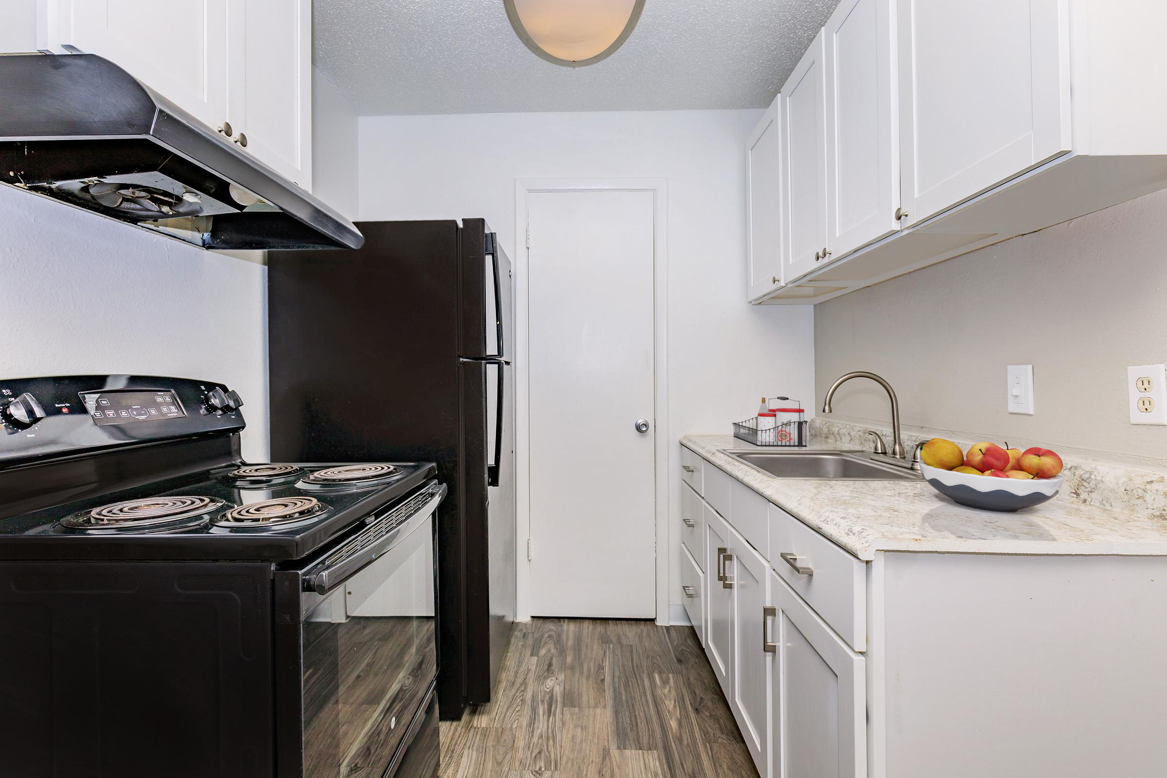 A modern kitchen featuring black appliances, including a stove and refrigerator, alongside white cabinets and a countertop with a bowl of colorful apples. The room has a light fixture above, wooden flooring, and a closed door visible in the background.