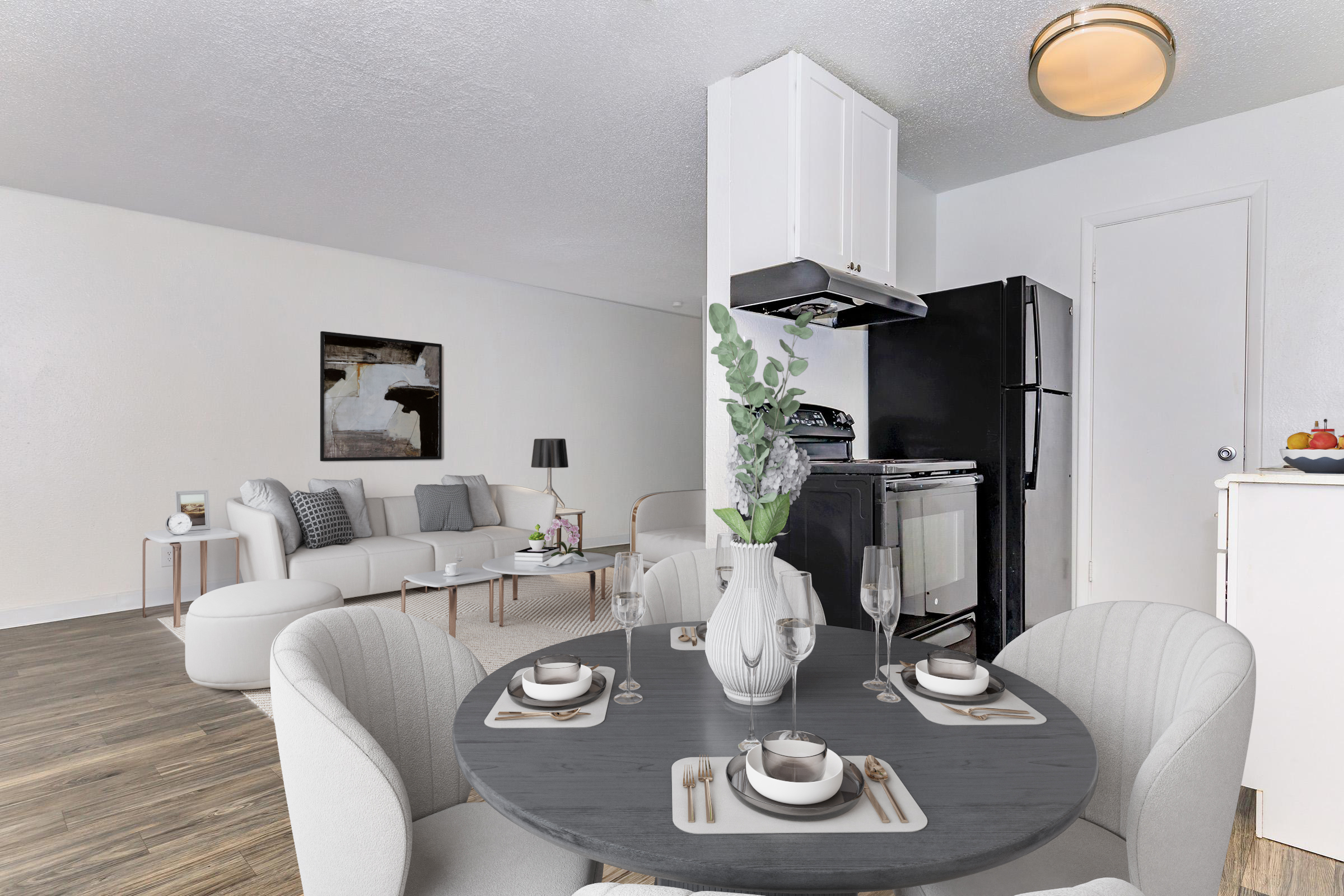 A modern living space featuring a dining area with a round table set for two, complete with plates, glasses, and utensils. In the background, there is a cozy seating area with a sofa and decorative artwork on the wall. The kitchen is visible, equipped with a black fridge and white cabinetry, all in a bright, airy atmosphere.