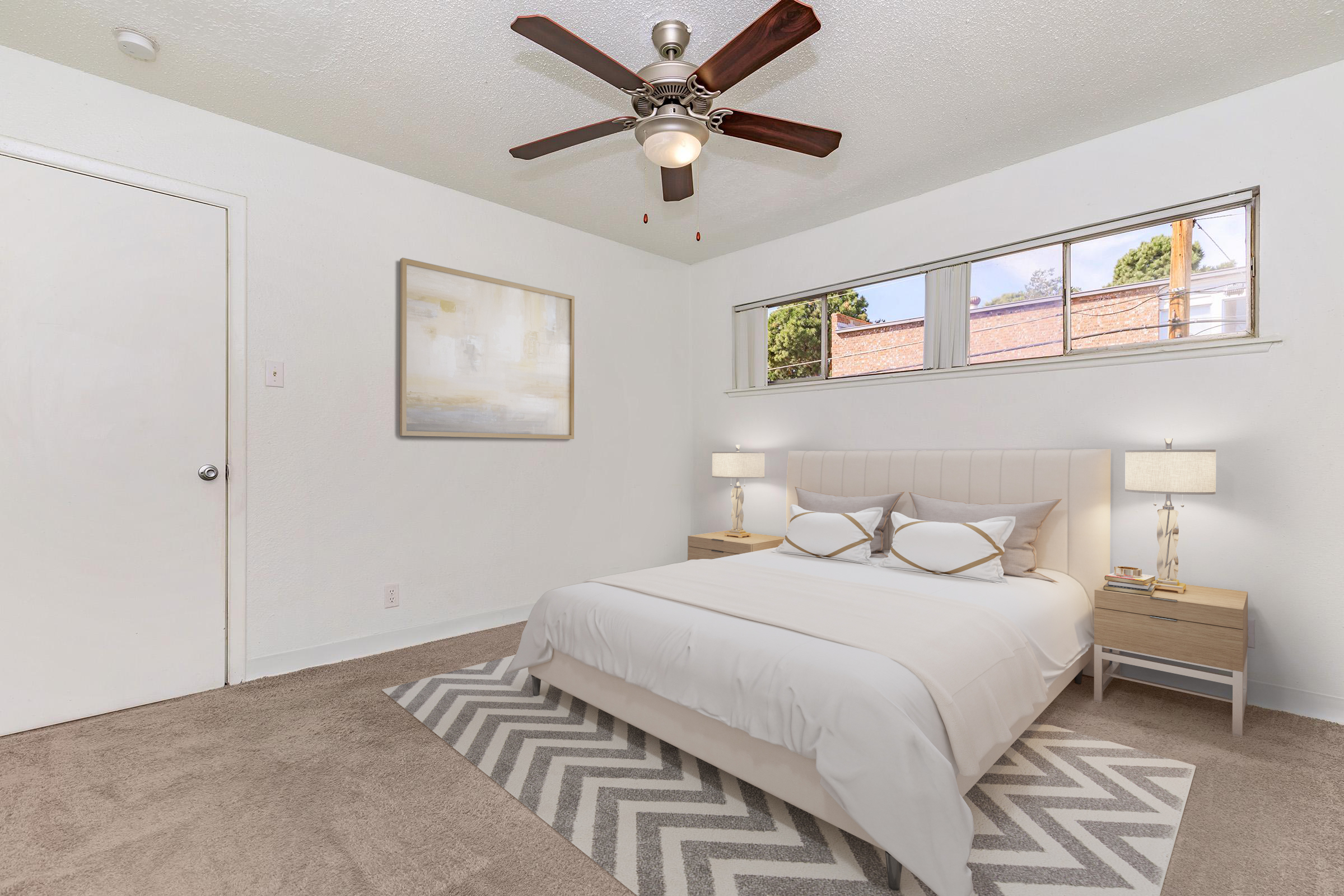 A cozy bedroom featuring a light-colored bed with decorative pillows, two nightstands, and lamps. A ceiling fan is visible, along with a large window letting in natural light. The walls are painted white, and a patterned rug lies on a carpeted floor. A piece of artwork is hung above the bed.