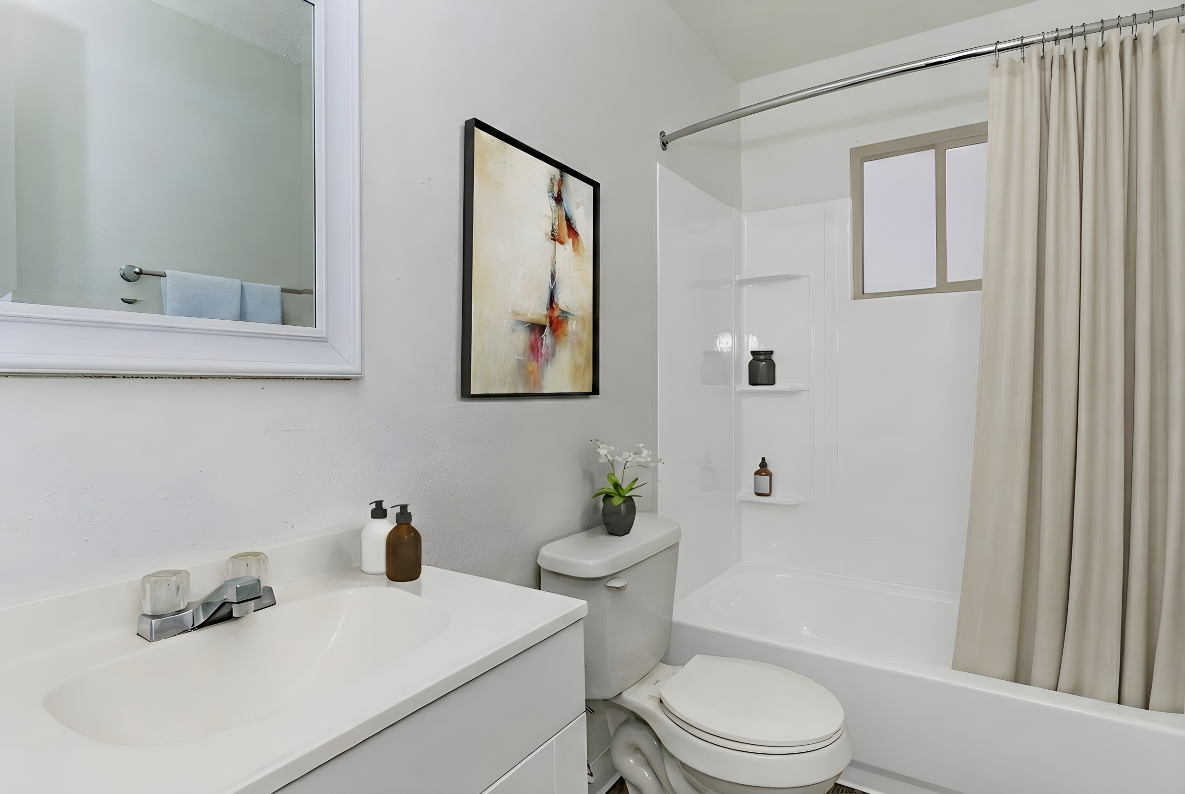 A modern bathroom featuring a white bathtub with a shower curtain, a vanity with a sink, and a round mirror. The walls are painted white, and there’s an abstract painting on the wall. There are decorative items on the vanity, including a small plant and various bottles. Soft lighting enhances the clean, minimalist aesthetic.
