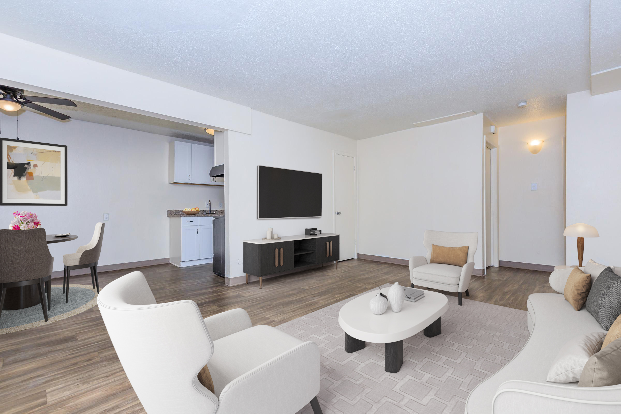 A spacious living area featuring two comfortable armchairs and a coffee table, with wooden flooring. In the background, a modern kitchen is visible, alongside a dining area with a round table. A large flat-screen TV is mounted on the wall, and there are soft, neutral colors throughout the room.