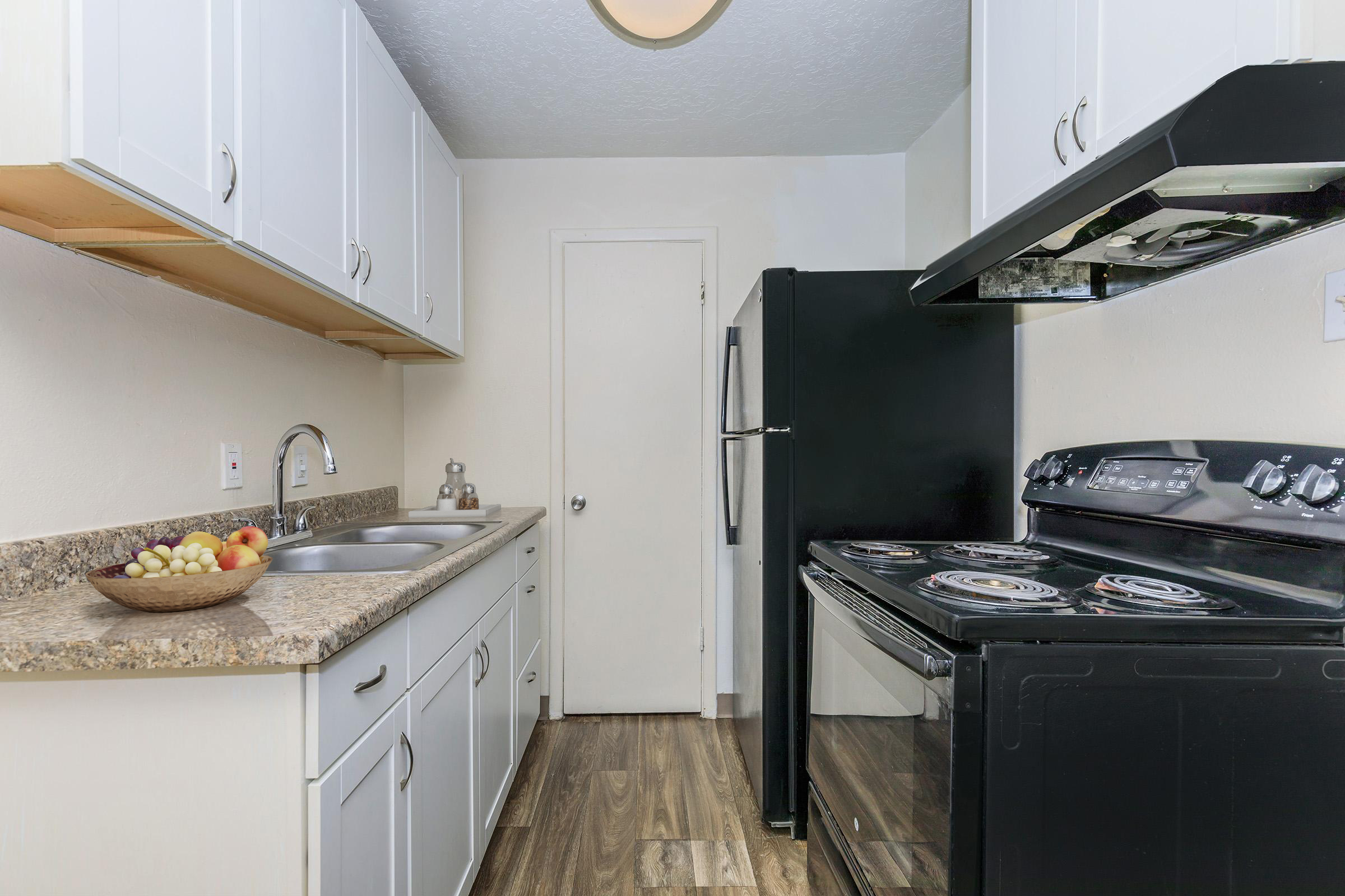 A modern kitchen featuring white cabinetry, a granite countertop with a bowl of fruit, a double sink, and black appliances including a refrigerator and an oven. The kitchen has wooden flooring and a door leading to another room. Natural light enters through the window above the sink.