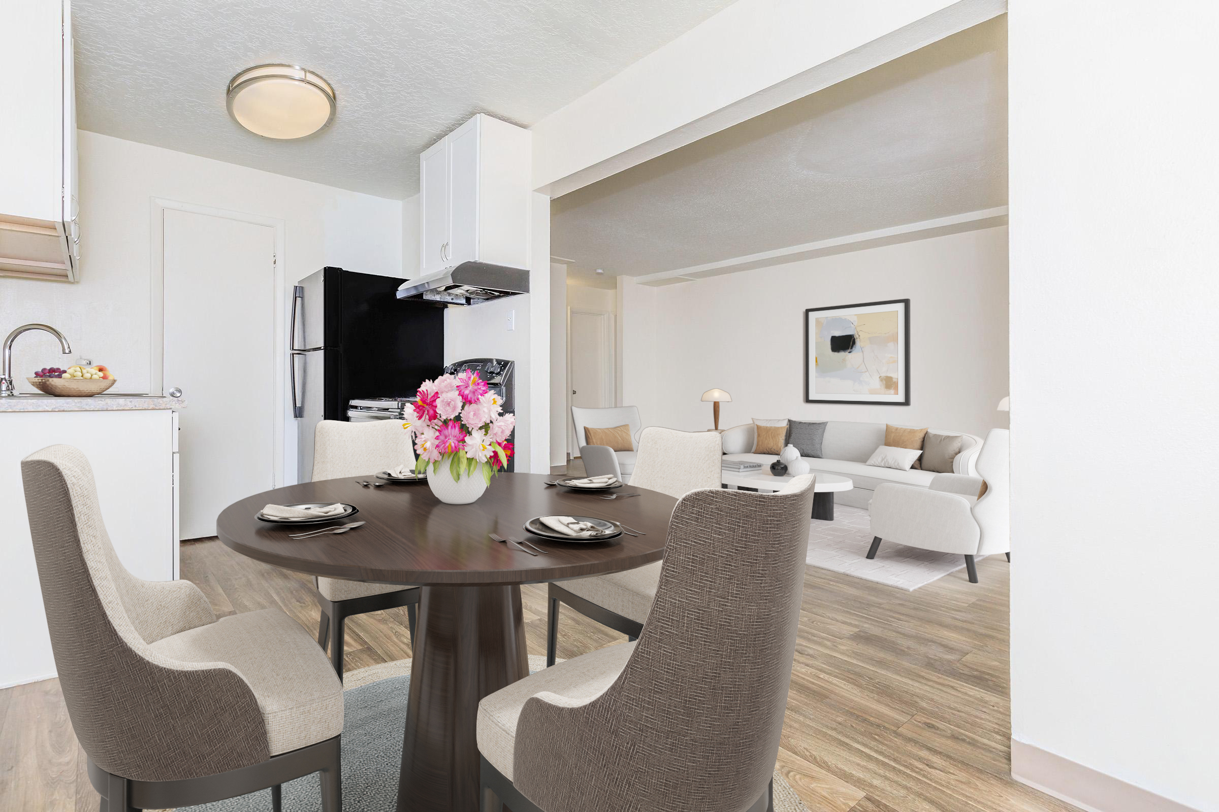 A modern dining area featuring a round table set with plates and a colorful flower centerpiece. Surrounding the table are four stylish chairs. In the background, a sleek kitchen is visible with a black refrigerator and light-colored cabinetry. A cozy living room with a sofa and artwork is beyond the dining space.