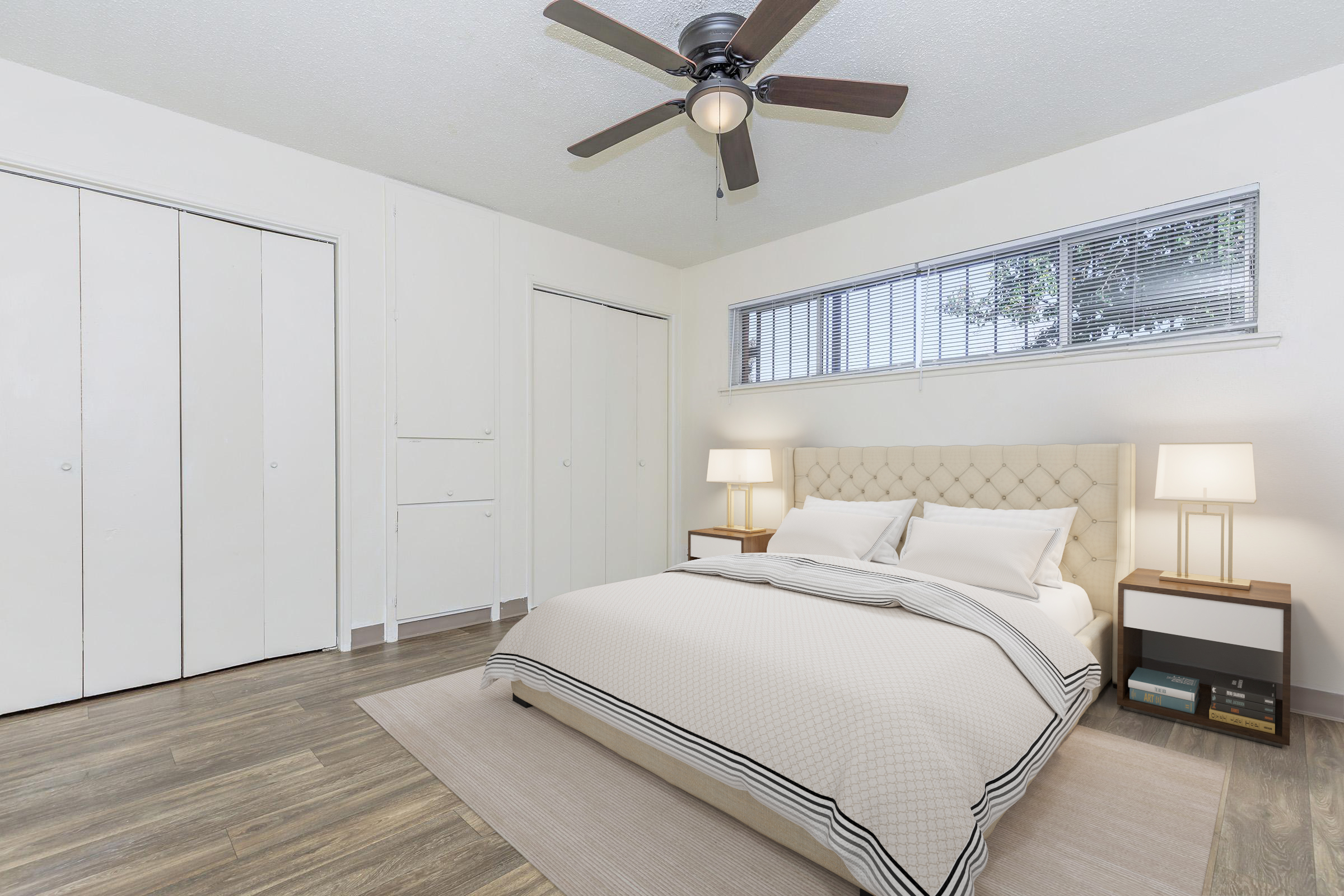 A bright and airy bedroom featuring a queen-sized bed with a beige tufted headboard and white bedding. There are two bedside tables with lamps, and a large window allowing natural light. The walls are light-colored, and the floor is covered with wood-like laminate. Closet doors are visible on one side of the room.