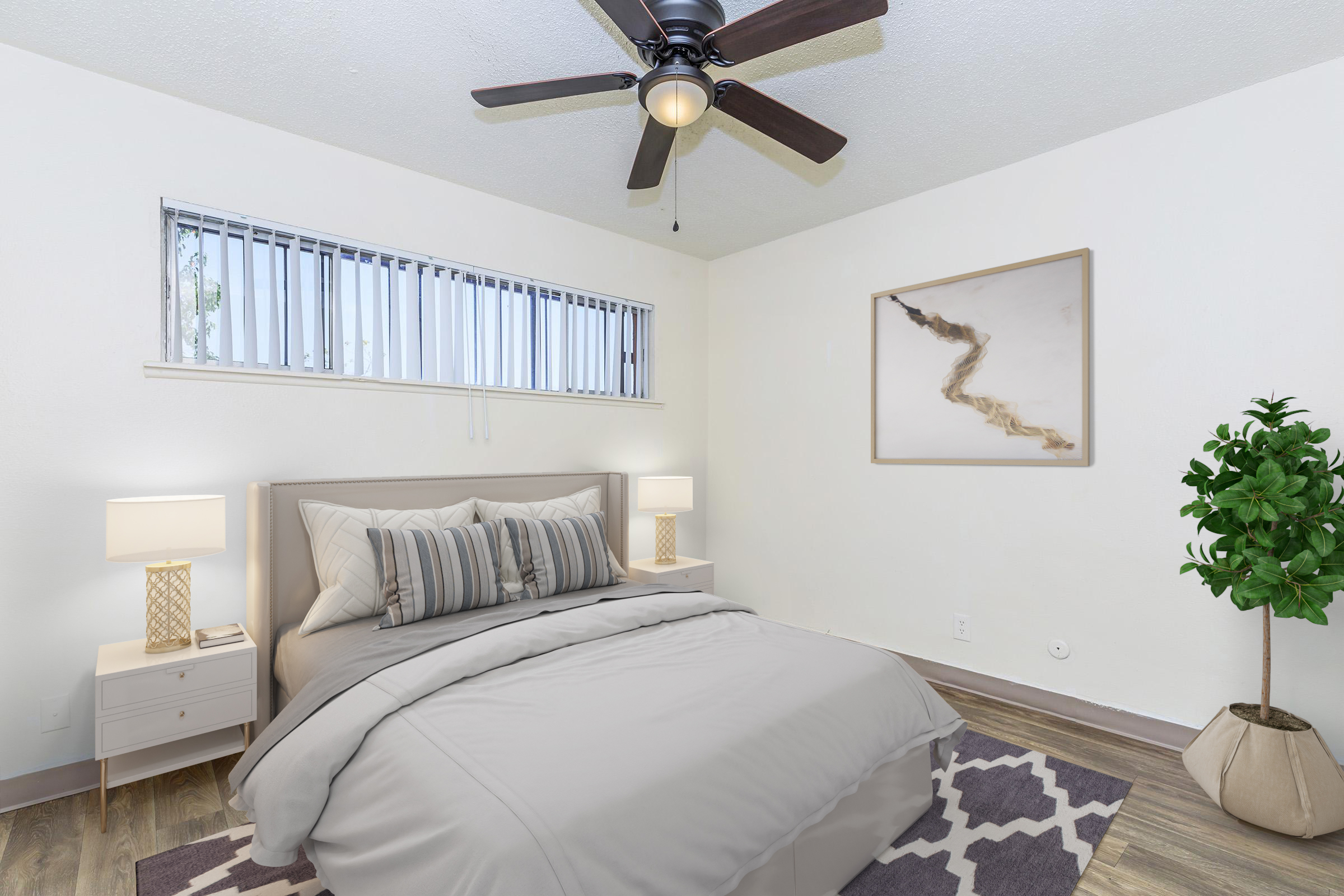 A cozy bedroom featuring a light-colored bed with gray bedding and decorative pillows. There are two bedside lamps on either side, a ceiling fan overhead, and a framed artwork on the wall. A potted plant adds a touch of greenery, while a patterned rug lies on the wooden floor. Natural light comes in through the window.
