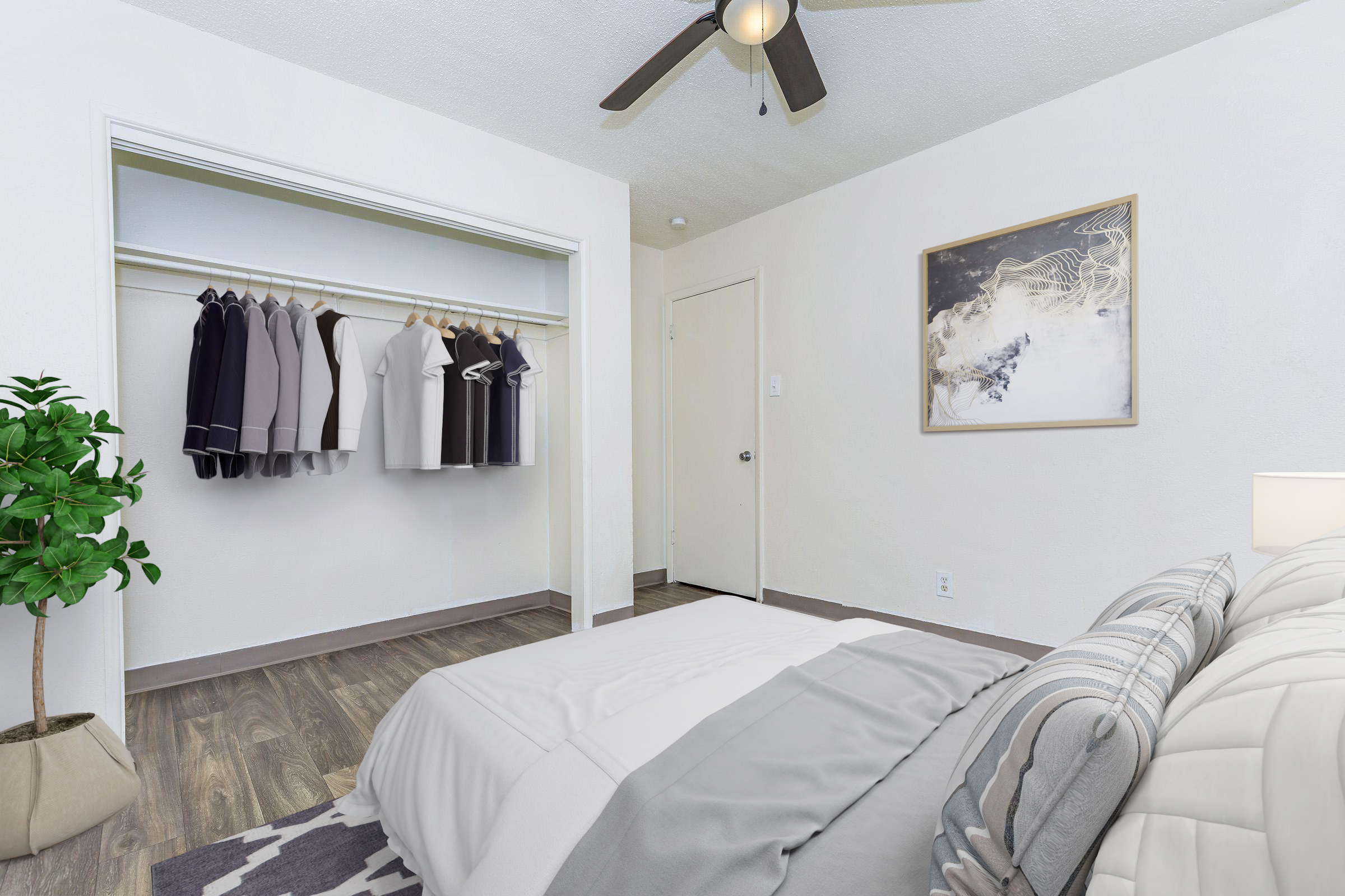 A neatly arranged bedroom featuring a bed with light gray bedding, a small bedside lamp, and a closet with neatly hung shirts. A potted plant adds a touch of greenery. The room has a light, airy feel with a ceiling fan and modern decor. The closet door is partially closed, leading to an inviting space.