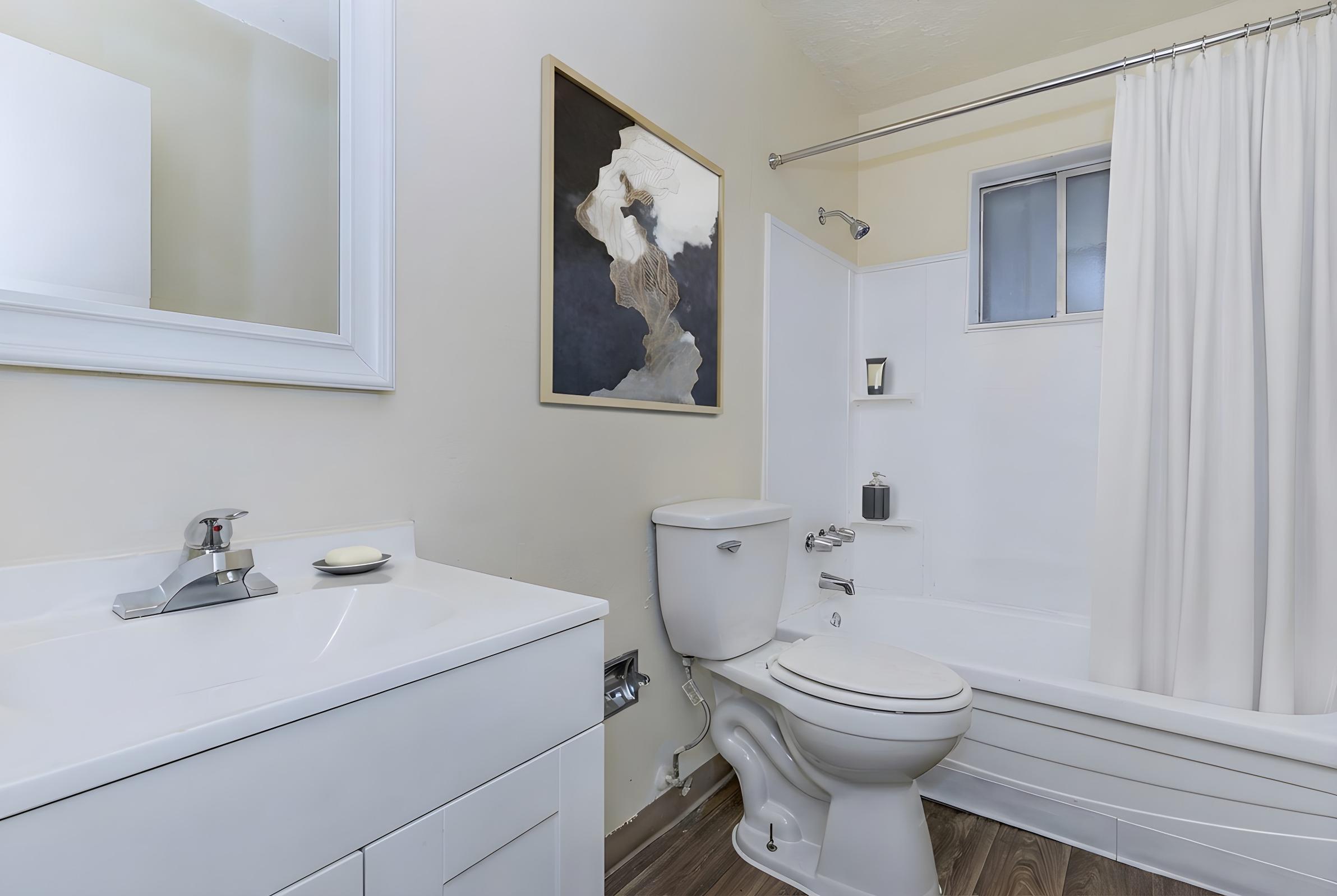 A clean and modern bathroom featuring a white bathtub with a shower curtain, a toilet, and a sleek vanity with a sink. The walls are painted in a light color, and there’s a piece of abstract art above the toilet. Natural light comes through a small window, and the flooring is a warm wood tone.
