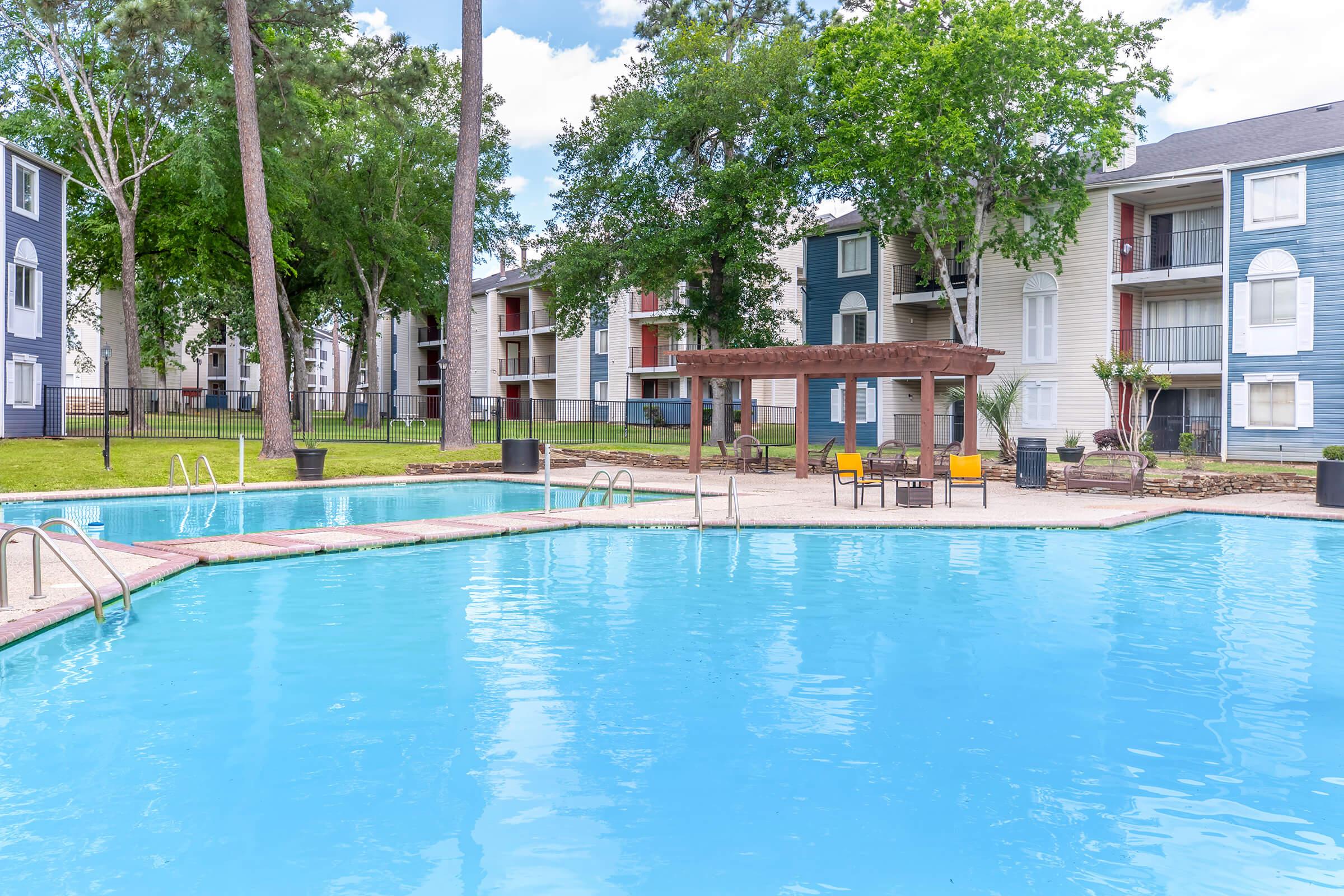 A serene swimming pool surrounded by green trees and shrubs, featuring lounge chairs under a wooden pergola. In the background, landscaped apartment buildings showcase a modern design, with clear blue skies above. The scene conveys a relaxing outdoor atmosphere, perfect for leisure and enjoyment.