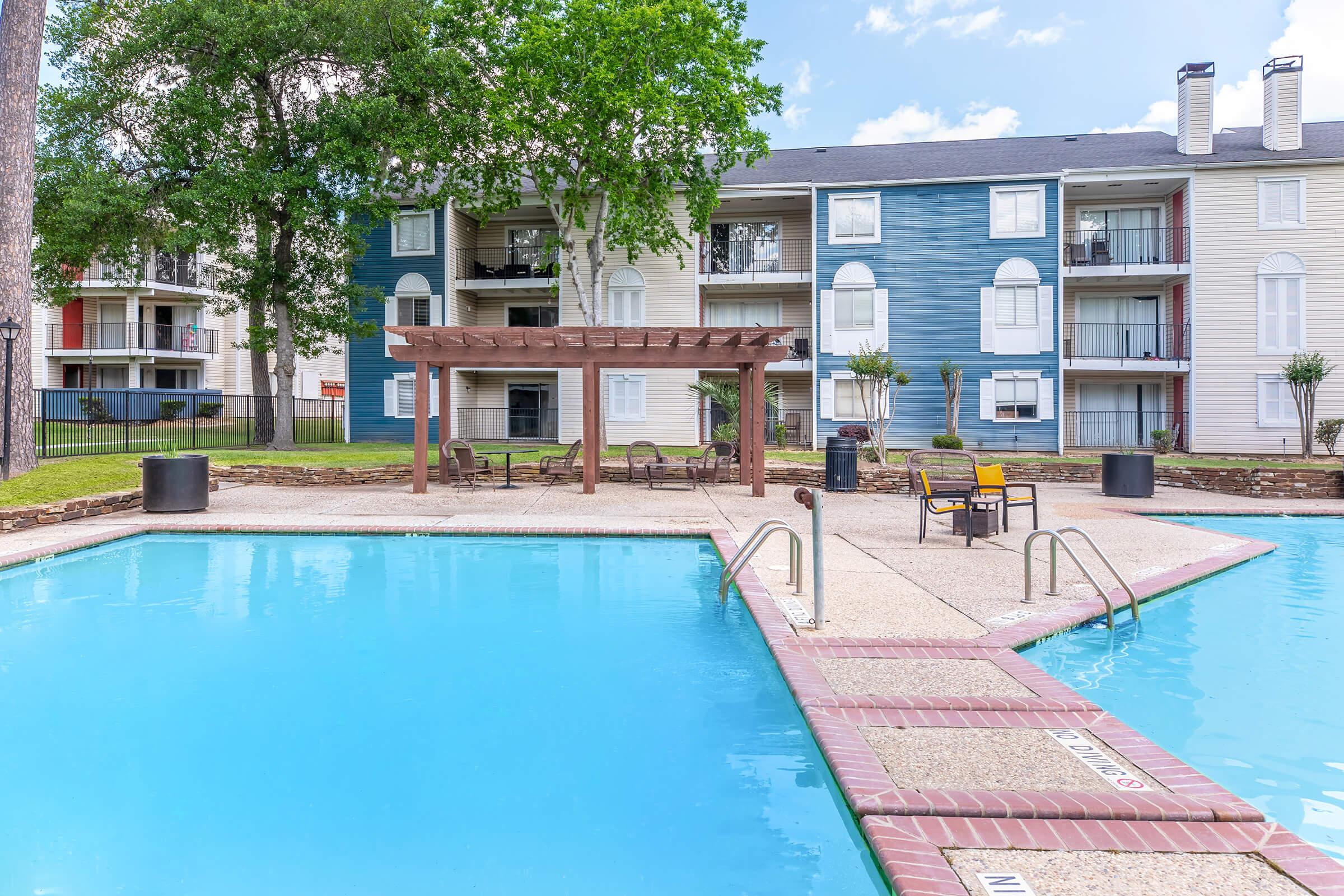 A sparkling blue swimming pool with a diving board and lounge chairs is surrounded by a landscaped area. In the background, a multi-story apartment building features balconies and is partially shaded by trees. The sky is clear with a few clouds, adding to the inviting atmosphere of the pool area.