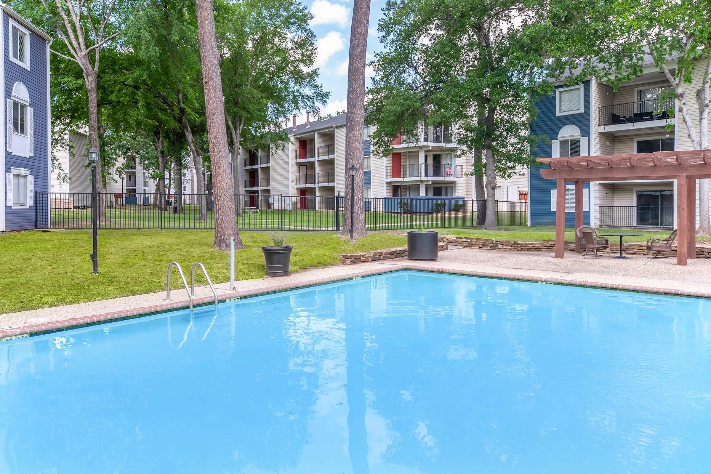 A clear blue swimming pool surrounded by green grass and palm trees. In the background, there are several apartment buildings with balconies, and a pergola with seating nearby. The scene is bright and inviting, suggesting a relaxing outdoor atmosphere.