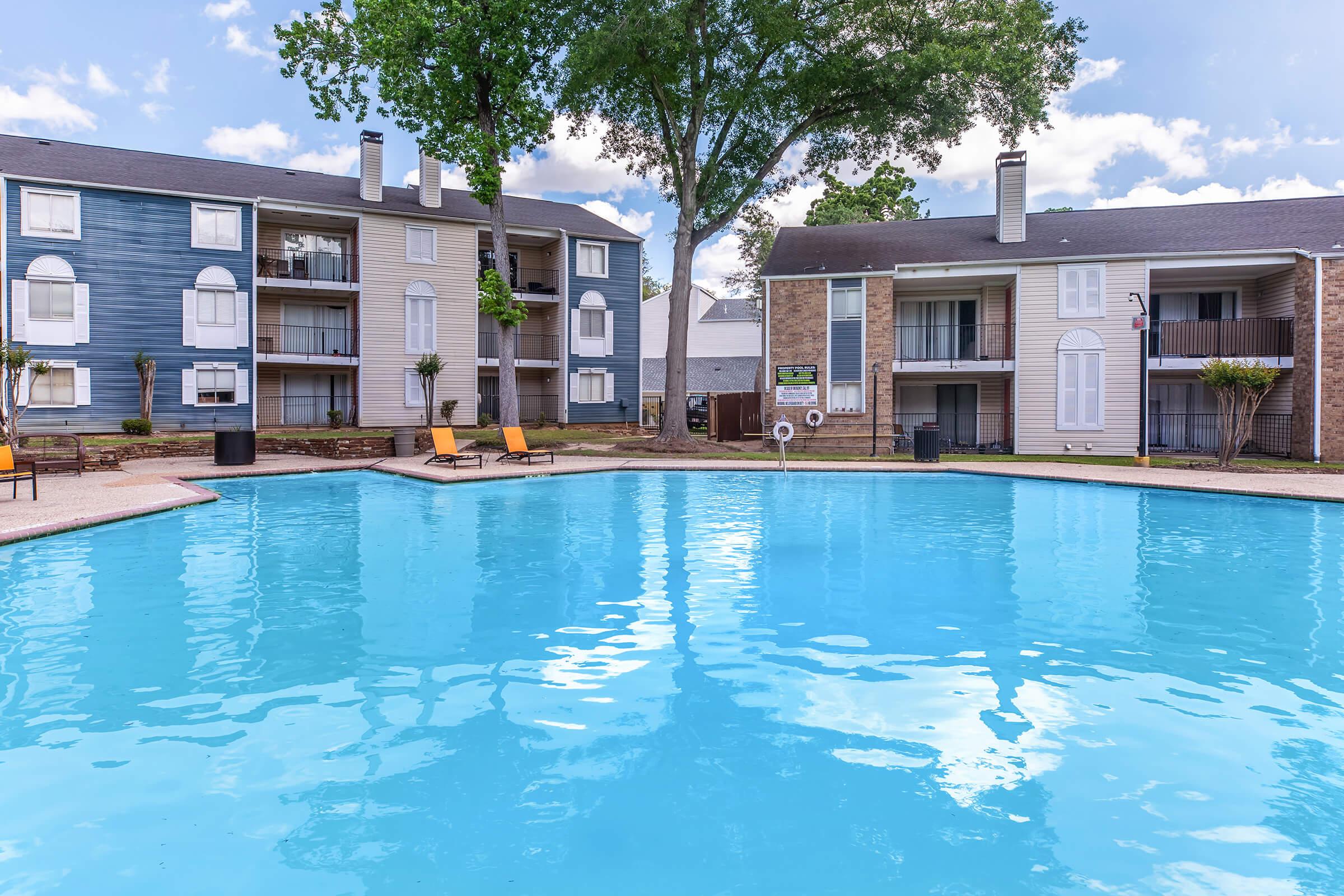 A serene swimming pool surrounded by two apartment buildings. The pool is clear blue, with lounge chairs on the deck. Lush trees are visible in the background, and the sky is partly cloudy. The overall atmosphere is relaxed and inviting, ideal for leisure or socializing.