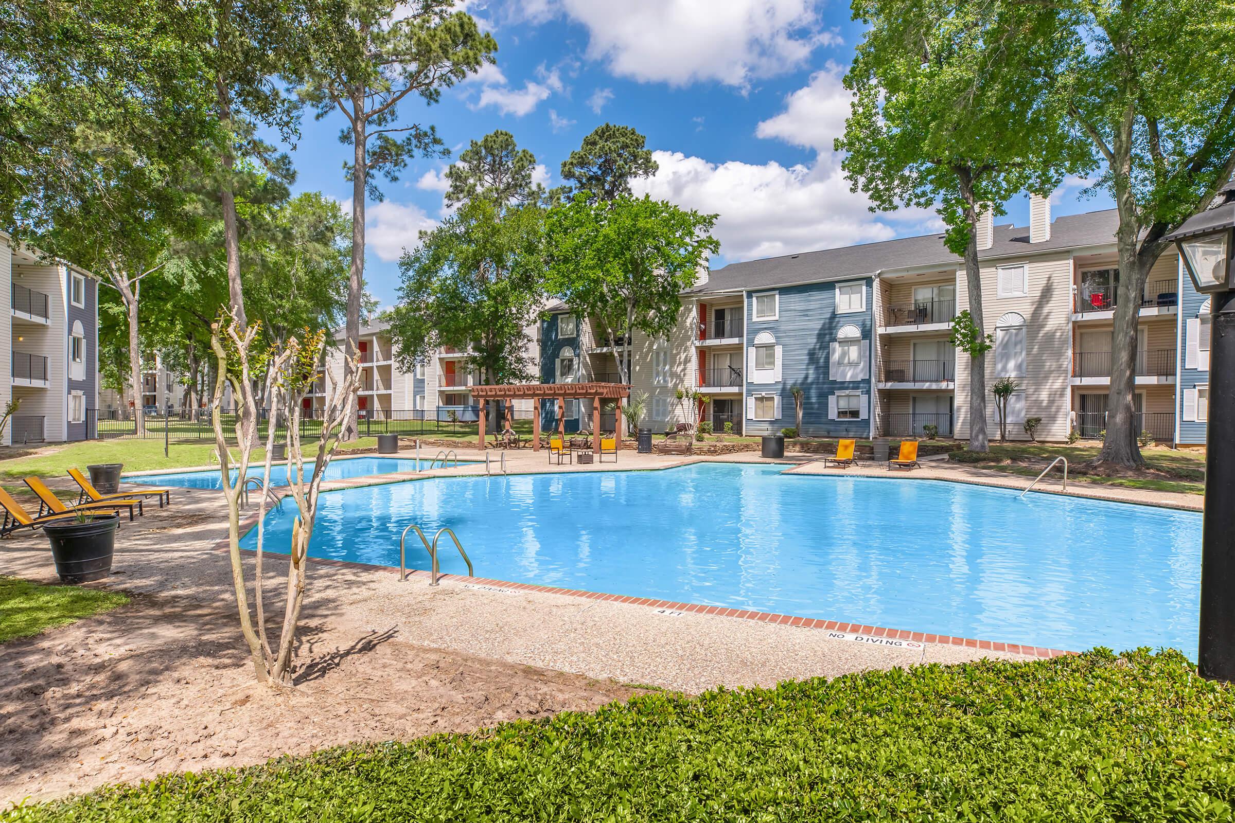 A clear blue swimming pool surrounded by lush green grass and trees. In the background, there are multi-story apartment buildings with balconies. A wooden gazebo and lounge chairs are positioned near the pool, creating a relaxing outdoor atmosphere. The sky is partly cloudy and bright.