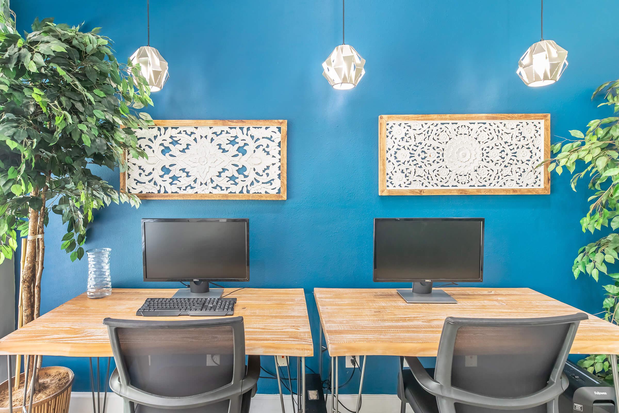 Two desks with black computer monitors and keyboards are positioned against a bright blue wall. Above the desks, there are two decorative framed artworks. Lush green plants are present on the sides, complemented by stylish hanging light fixtures. The overall space appears modern and inviting.