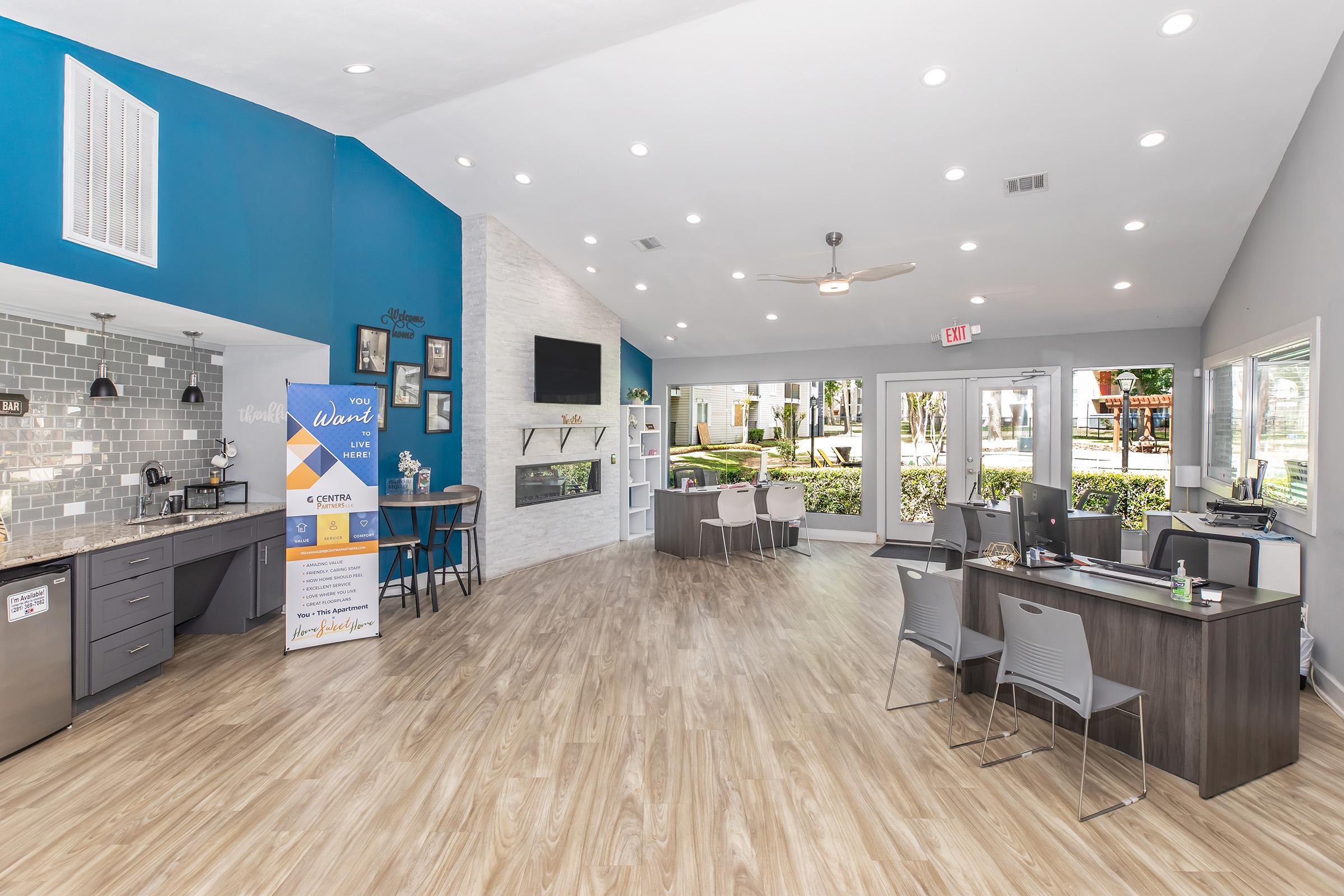 Interior view of a modern, brightly lit common area featuring a seating arrangement with tables and chairs. A kitchenette with gray cabinets is on the left, while a TV and fireplace are on a white stone wall. Large windows offer a view of outdoor greenery, with a blue accent wall enhancing the ambiance.