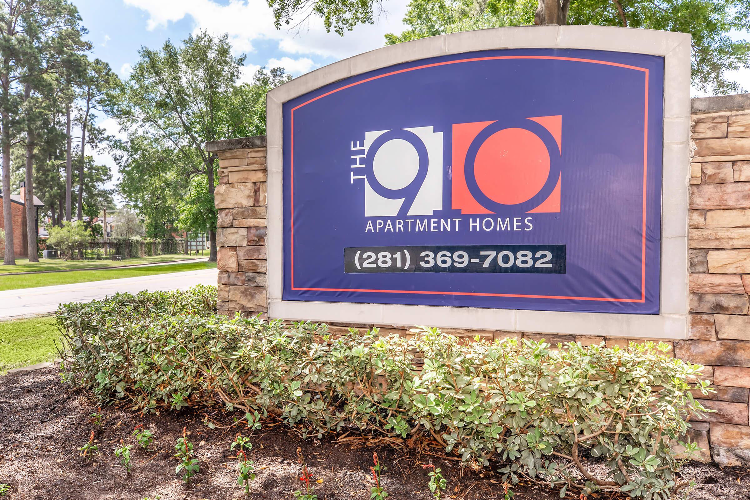Sign for The 910 Apartment Homes, featuring a prominent logo with the number 910 and the text "Apartment Homes." The sign is surrounded by landscaping, including shrubs. A phone number, (281) 369-7082, is displayed for inquiries. The setting includes trees and a driveway in the background.