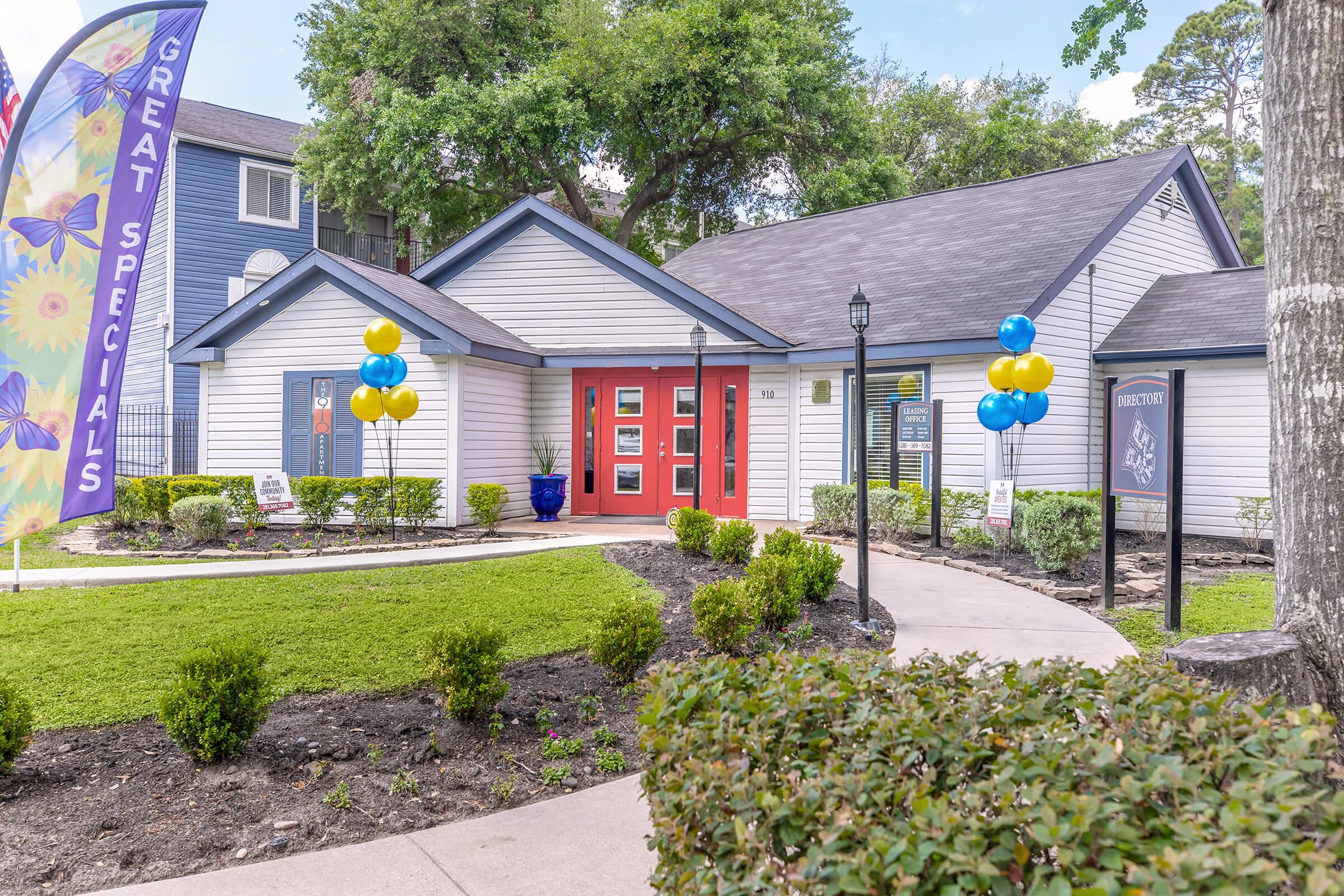 A well-maintained building with a gray exterior and red doors, surrounded by neatly landscaped greenery. Two decorative balloons in blue and yellow stand at the entrance. A colorful banner promotes great specials. A pathway leads to the front entrance, enhancing the inviting atmosphere of the location.