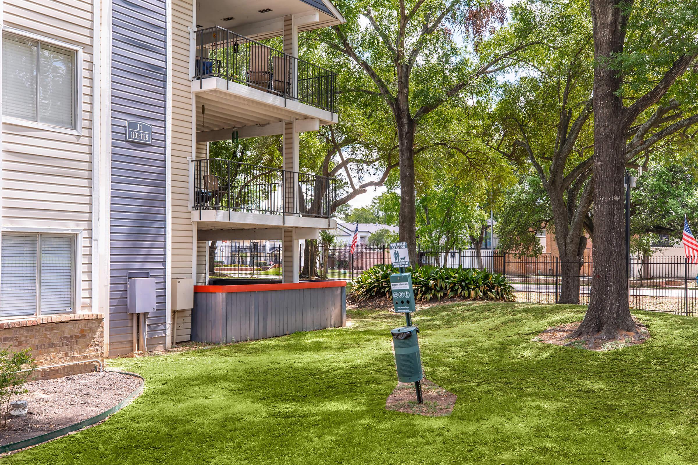 View of a well-maintained apartment complex featuring multiple balconies on two stories, surrounded by lush green grass and trees. A trash and recycling station is visible in the foreground, along with a fenced area and American flag in the background, indicating a residential environment.