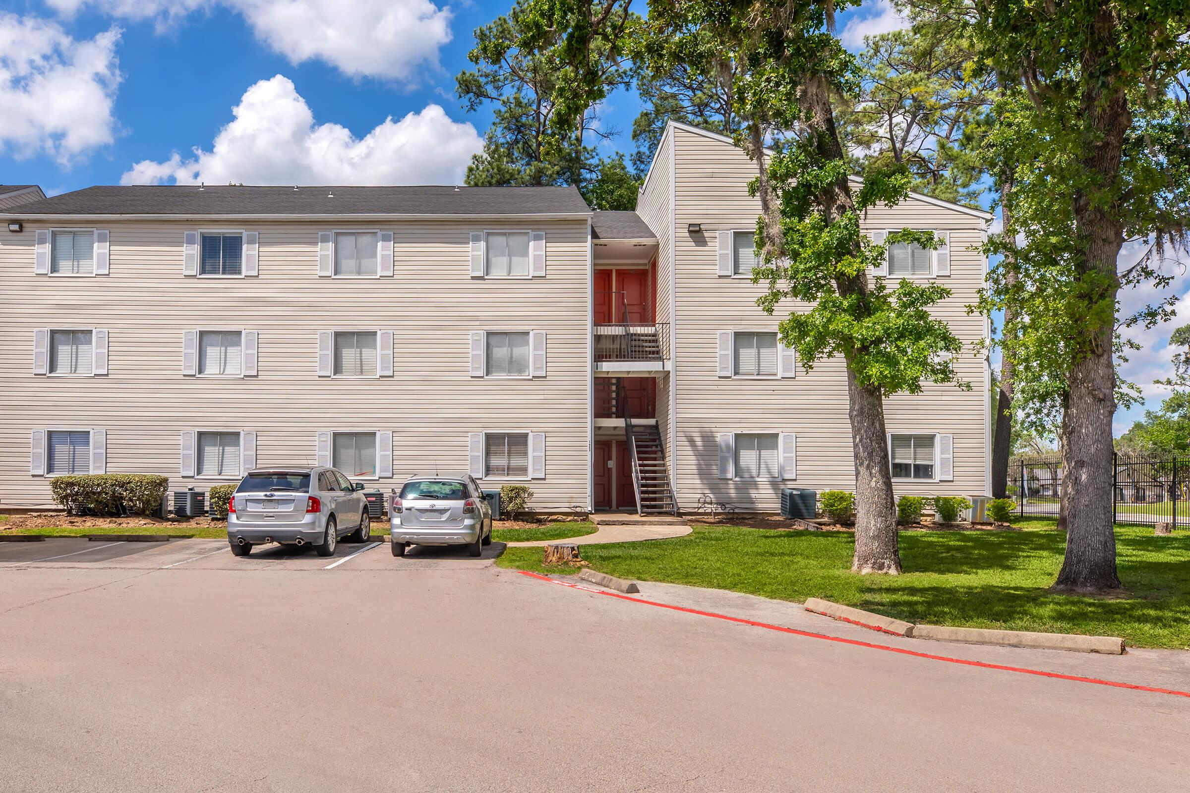 A three-story apartment building with light-colored siding, featuring several windows and a central staircase. The surrounding area has parking spaces for vehicles, including two parked cars. Lush green grass and trees are visible in the foreground, under a partly cloudy blue sky.