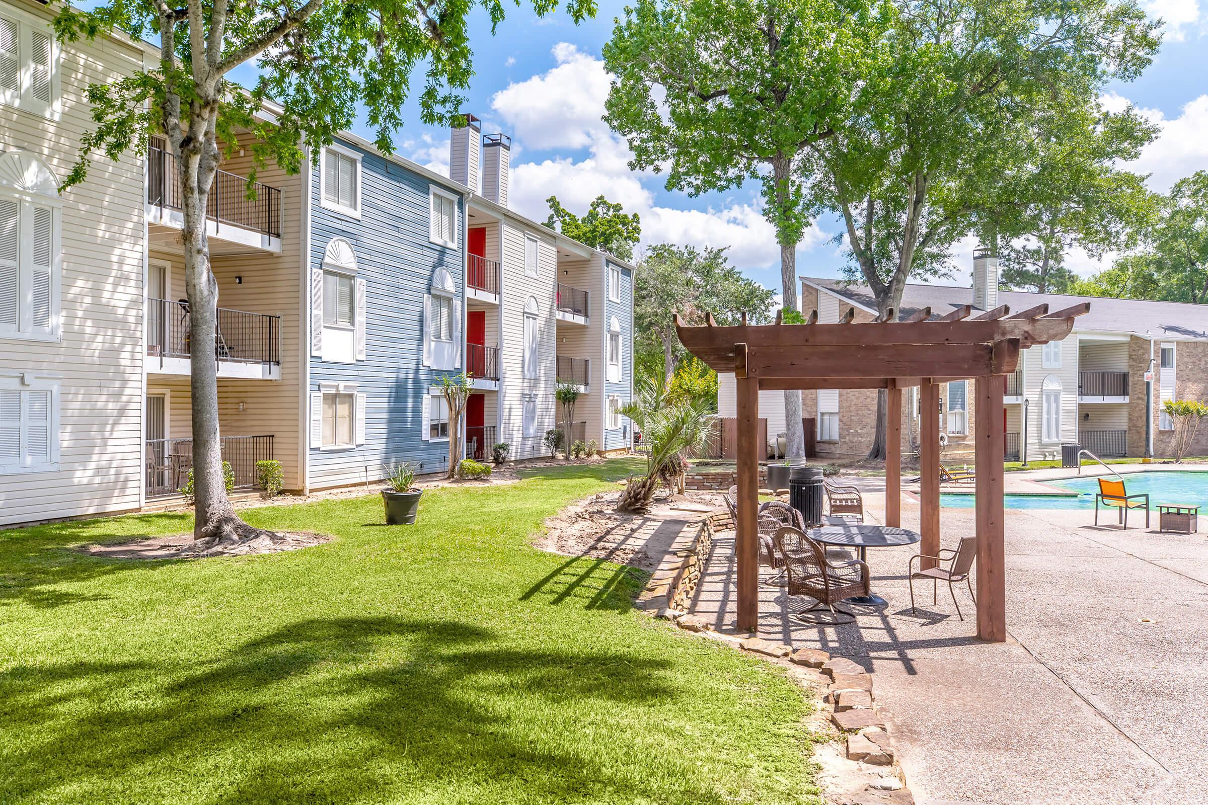 A well-kept outdoor area of an apartment complex featuring green lawns, trees, and a pergola with seating. In the background, several apartment buildings are visible, along with a swimming pool. The scene is set under a clear blue sky with scattered clouds, creating a pleasant and inviting atmosphere.