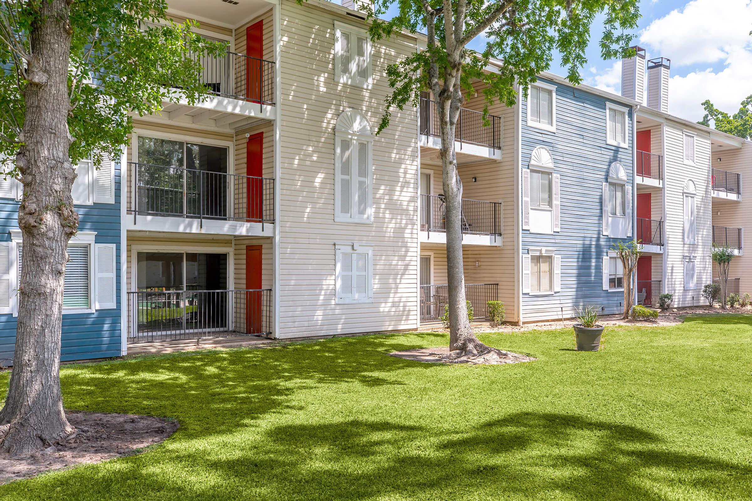 A multi-story apartment building featuring a mix of blue and beige exterior walls. The building has balconies with railings and large windows. Lush green grass surrounds the building, complemented by trees in the foreground, creating a serene outdoor space.