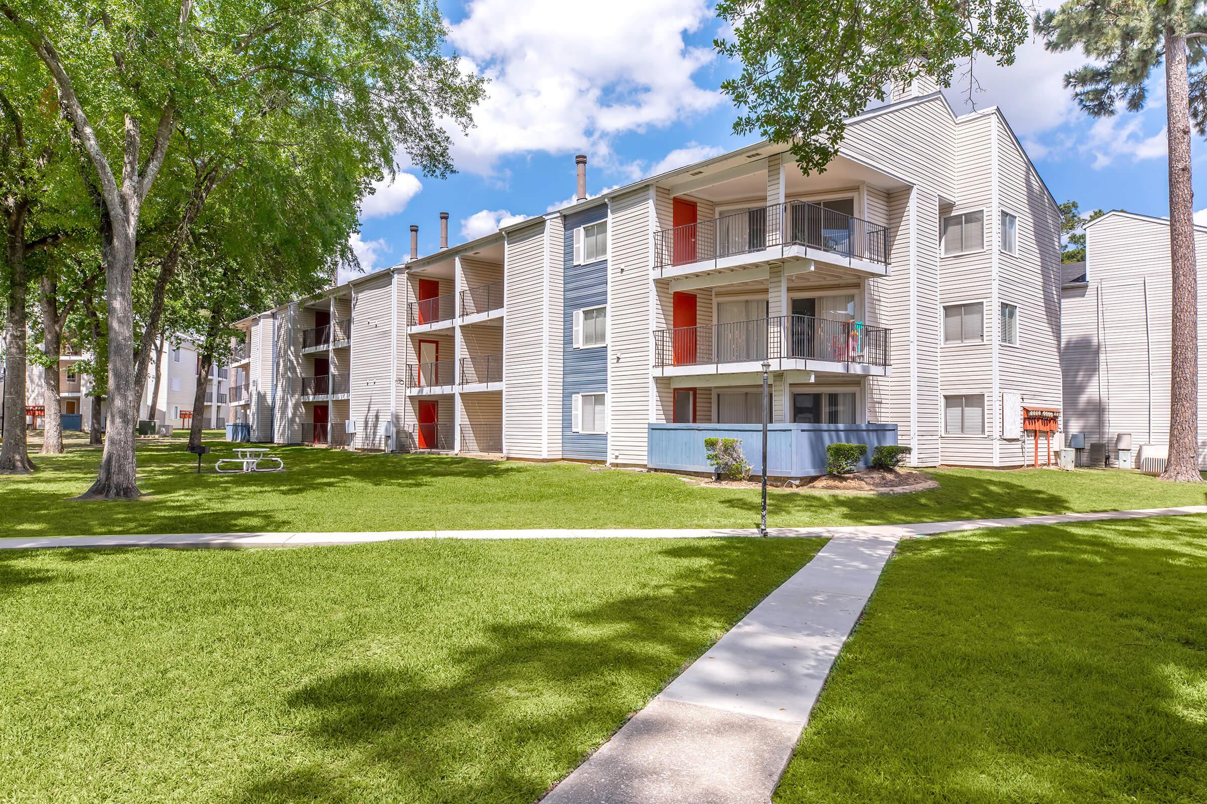 A well-maintained apartment complex with multiple buildings featuring balconies and red doors. The surrounding landscape includes lush green grass and trees, with a concrete pathway leading through the lawn. The sky is partly cloudy, creating a bright and inviting atmosphere.