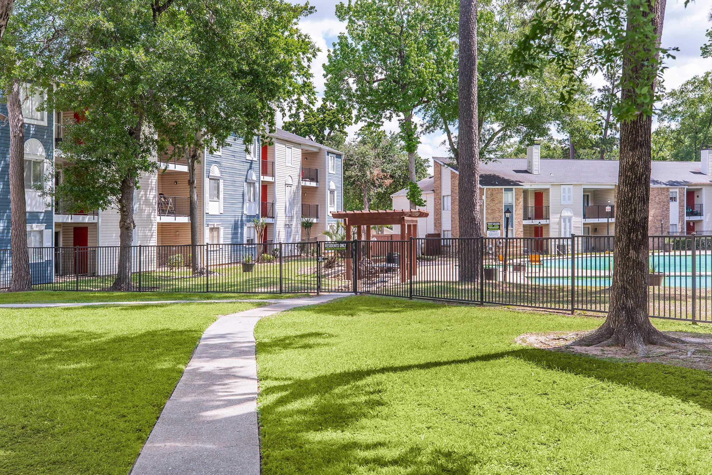 A well-maintained apartment complex with two-story buildings, surrounded by lush green grass and trees. A walkway leads through the landscape towards a fenced pool area with lounge seating and a shaded pergola, creating a welcoming outdoor space for residents. Clear blue skies are overhead.