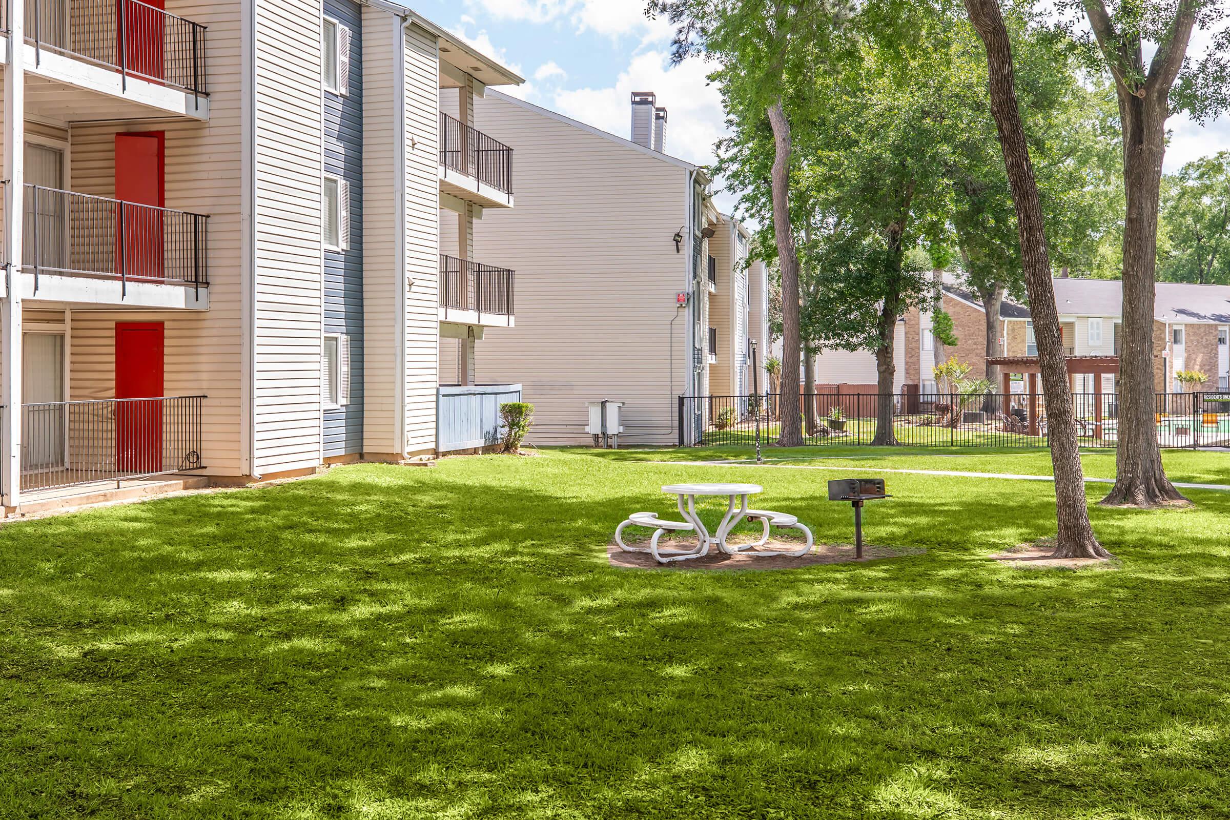 A well-maintained grassy area with a picnic table and grill surrounded by apartment buildings. The scene features bright green grass, trees providing shade, and a clear sky, creating a welcoming outdoor space for residents.