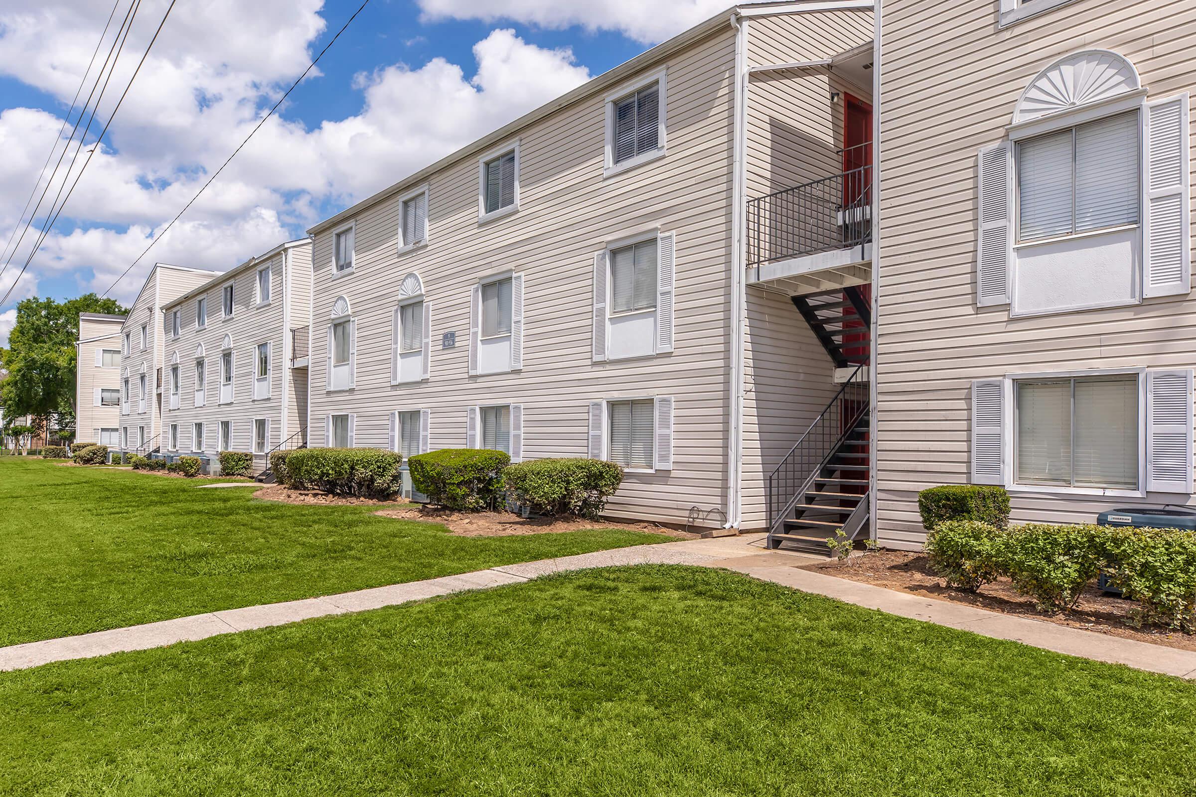 A view of a three-story apartment building with beige siding and multiple windows. Green grass and neatly trimmed bushes are in the foreground, and a set of stairs leads to a second-floor entrance. The sky is partly cloudy, adding to the bright and inviting atmosphere.