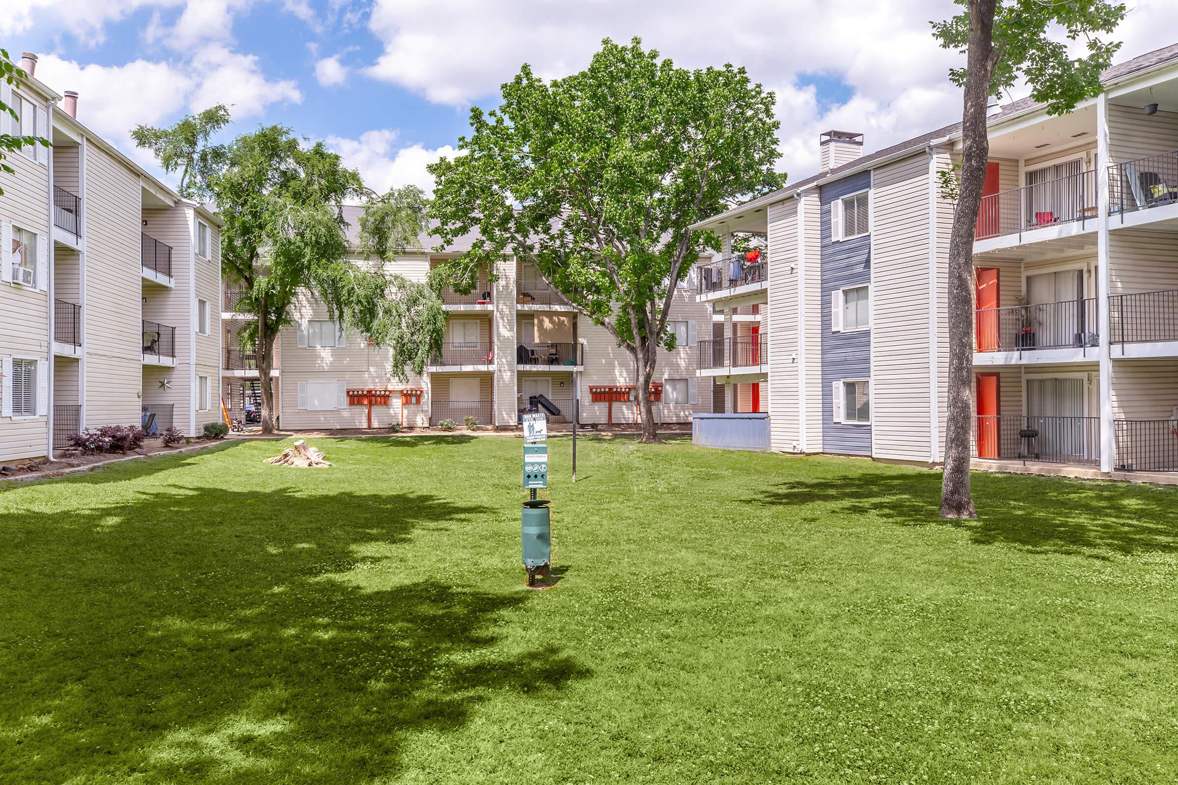 A well-maintained outdoor area featuring a green lawn surrounded by residential apartment buildings, with trees providing shade. There is a water fountain or feature in the center of the lawn, and balconies can be seen on the buildings. The sky is partly cloudy, creating a bright and inviting atmosphere.