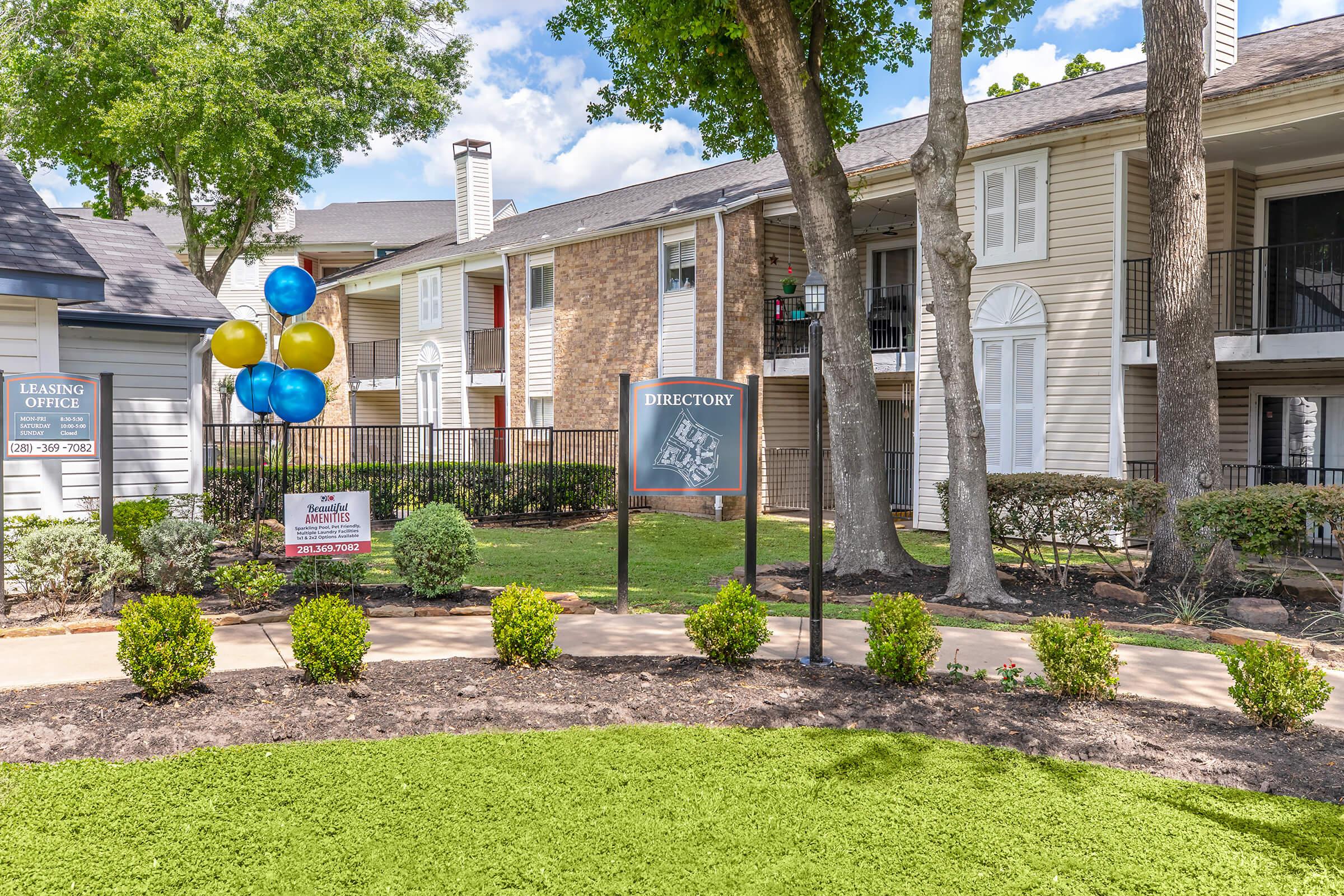A well-maintained apartment complex entrance featuring a sign for "Directory" surrounded by green landscaping. Colorful balloons in blue, yellow, and green are tied to a nearby fence. The leasing office is visible on the left, with several trees providing shade. Cloudy skies are overhead, creating a bright atmosphere.