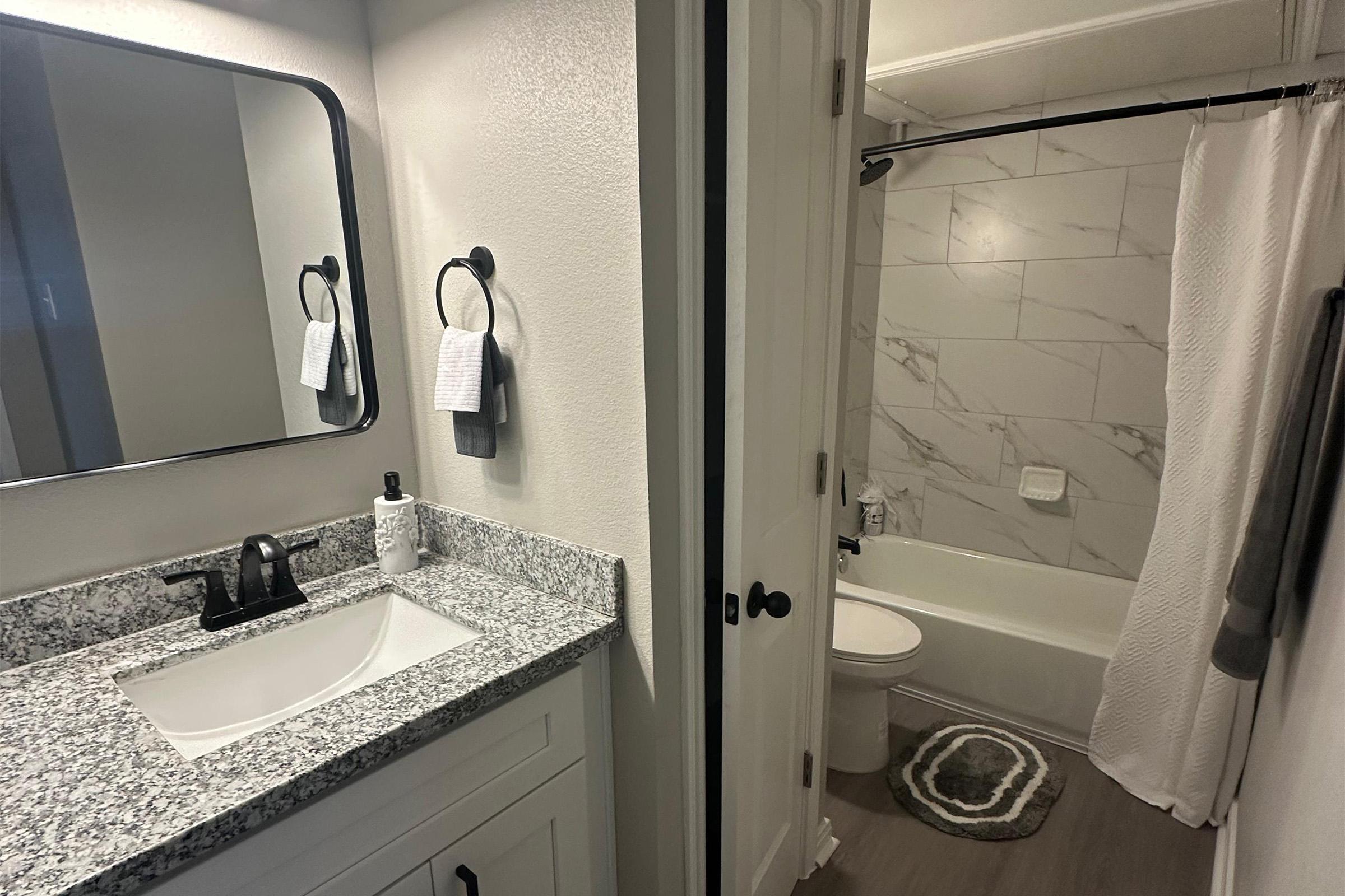 A clean and modern bathroom featuring a white vanity with a granite countertop and a black faucet. A large mirror hangs above the sink, with two towel holders beside it. On the right, there is a bathtub with a white shower curtain and marble-style tiles. The flooring is light-colored wood.
