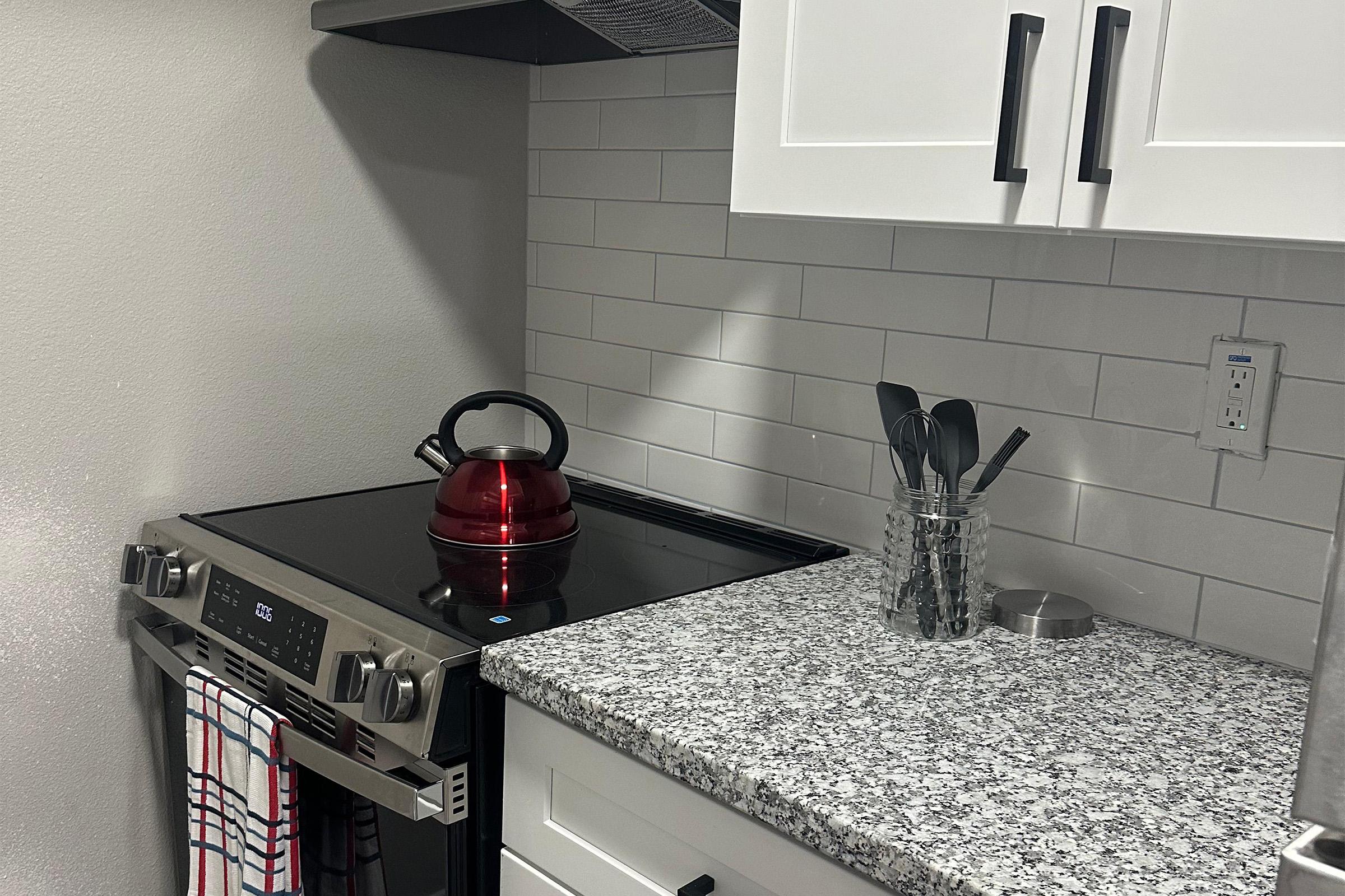 A modern kitchen corner featuring a black stovetop with a digital display, a red kettle on the burner, and a silver utensil holder filled with cooking tools. The countertop is made of speckled granite, and the white cabinets have black handles. A dish towel hangs from the stove.