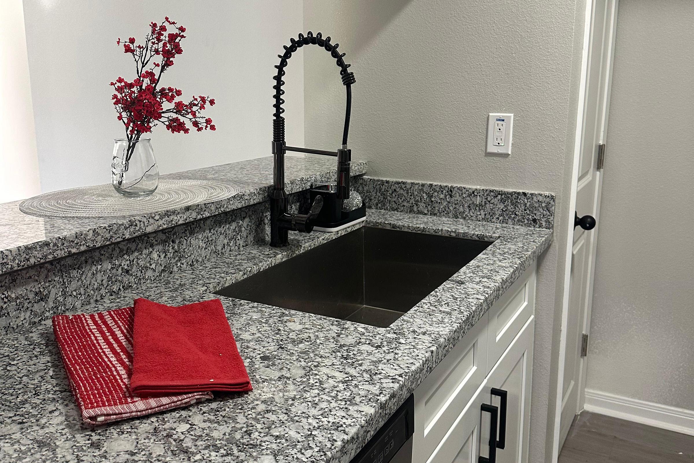 A modern kitchen countertop features a stainless steel sink with a sleek black faucet. A vase with decorative red flowers is placed on the counter, alongside a round placemat. Two red and white dish towels are neatly folded. The wall is light-colored, and a door is visible in the background.