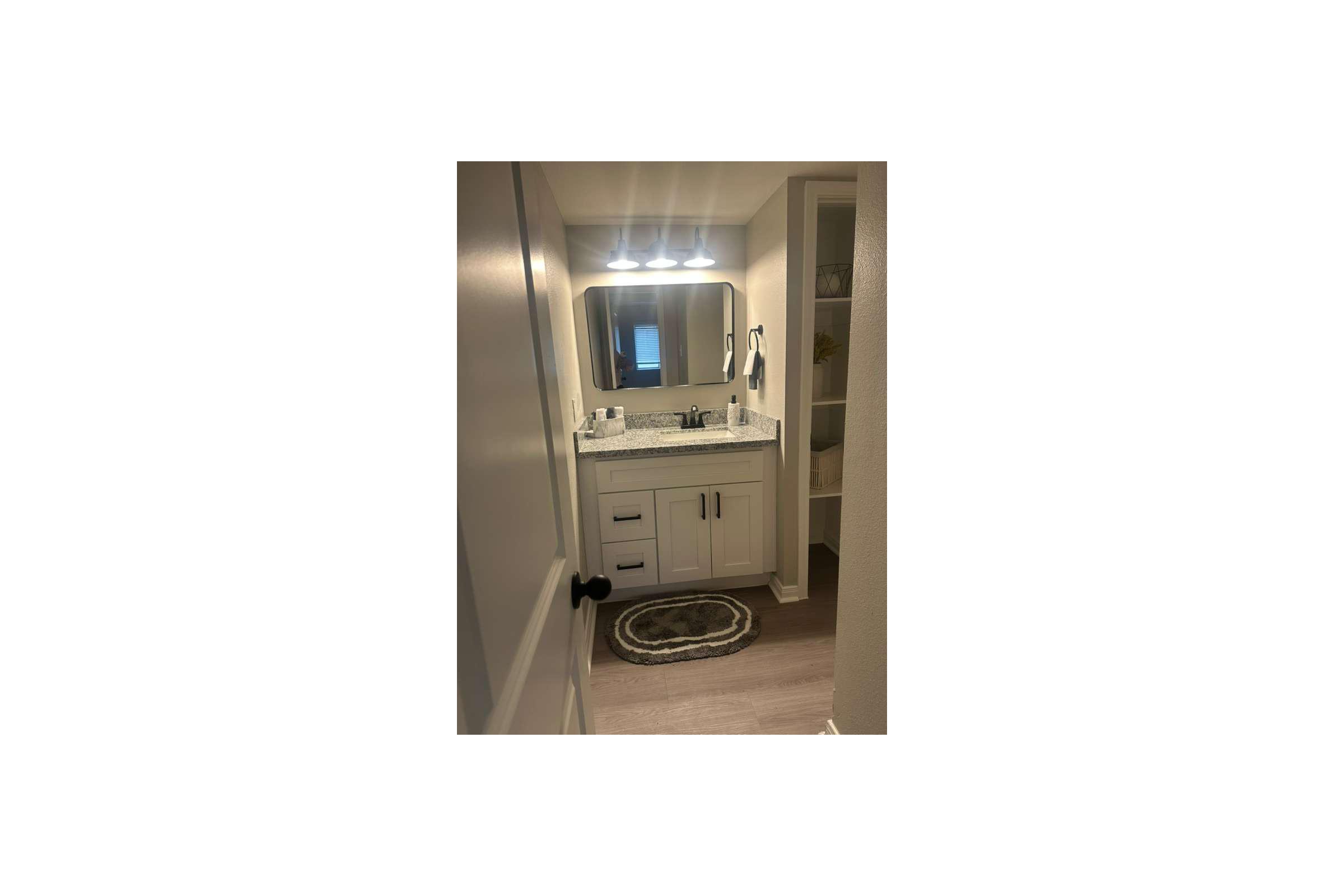 A view of a modern bathroom featuring a white vanity with a granite countertop, a large mirror with bright lighting above, and a round rug on the floor. The walls are painted a light color, and a door is visible on the left side of the image. The overall design is clean and contemporary.