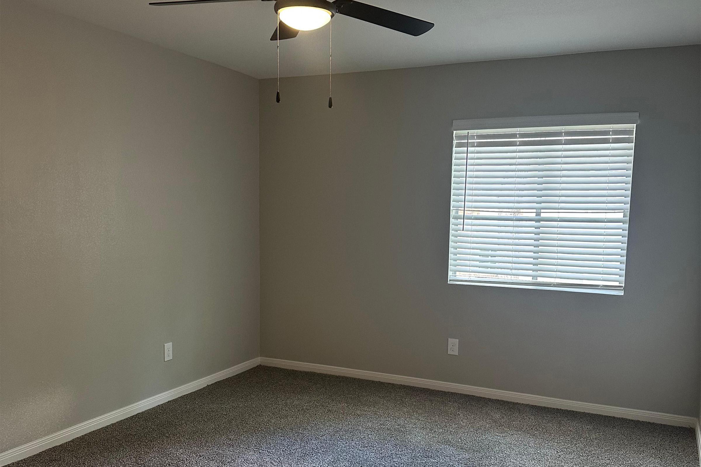A blank room with light gray walls and carpeted flooring. A ceiling fan with lights is mounted on the ceiling, and there is a window with white blinds allowing natural light to enter. The room appears empty and unfurnished, creating a spacious and neutral ambiance.