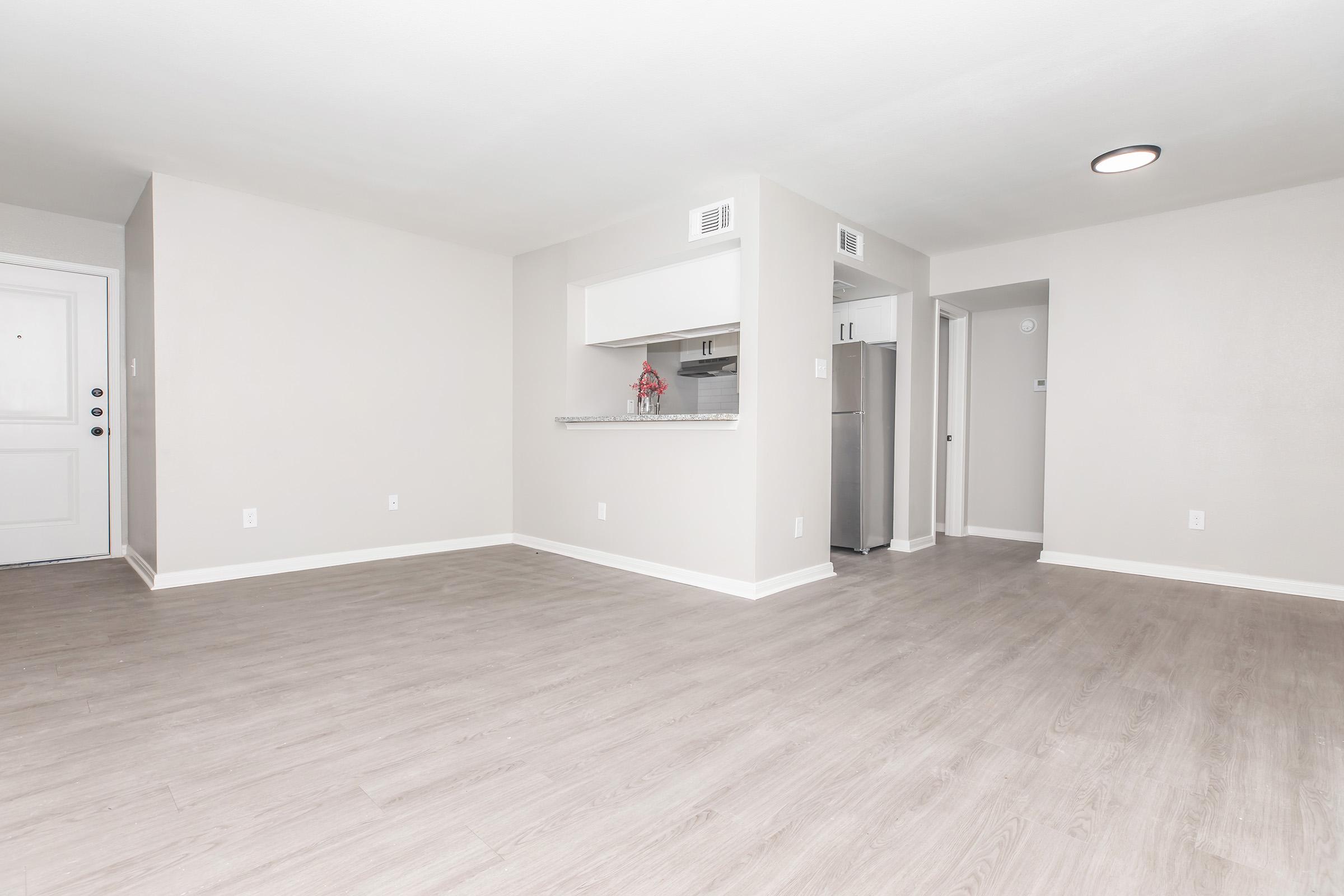 Spacious, modern living room with light gray walls and new flooring. Features an open layout leading to a kitchen area with a bar, stainless steel refrigerator visible. The entrance door is on the left, and there is a hallway leading to another room, creating a bright and inviting atmosphere.