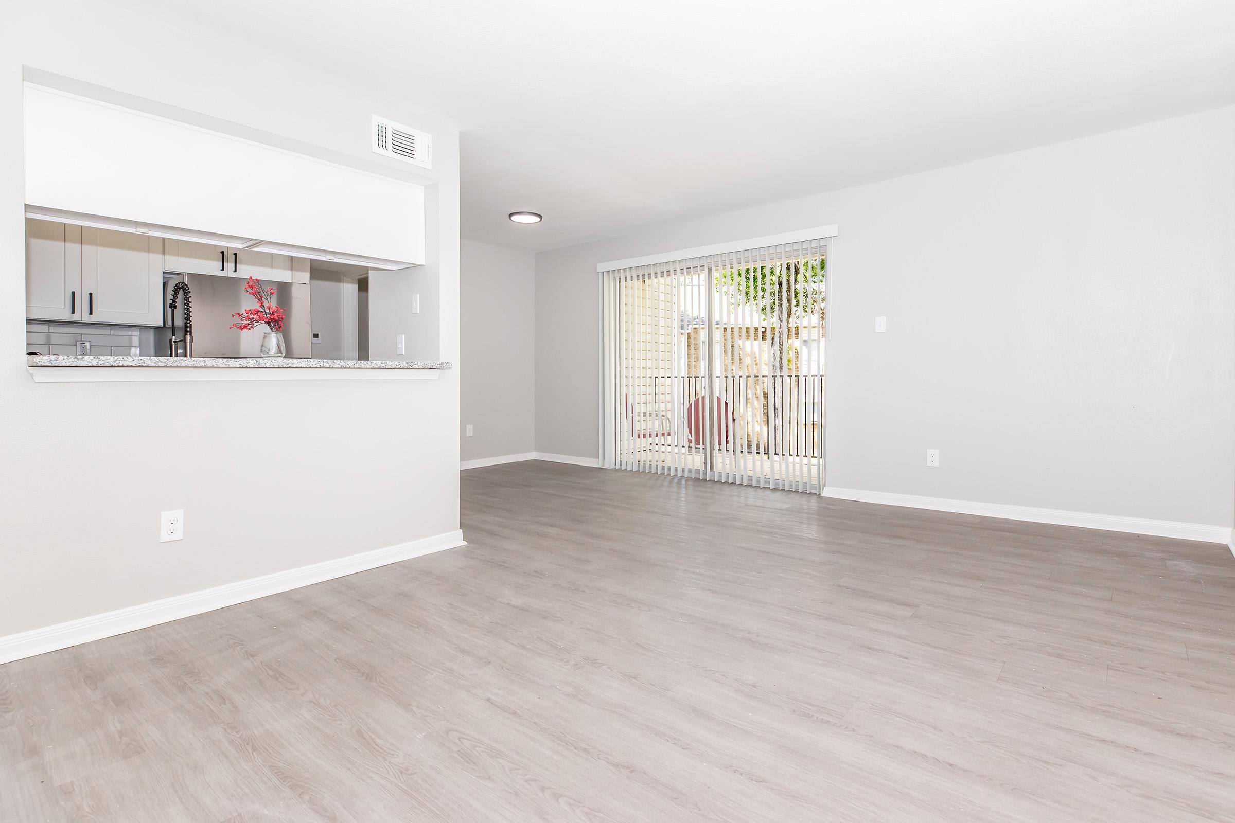 Spacious interior of a modern living area featuring light-colored walls, vinyl flooring, and an open layout. A sliding glass door leads to an outdoor space, and a kitchen is visible in the background with a breakfast bar. Bright and inviting atmosphere with natural light filtering in.