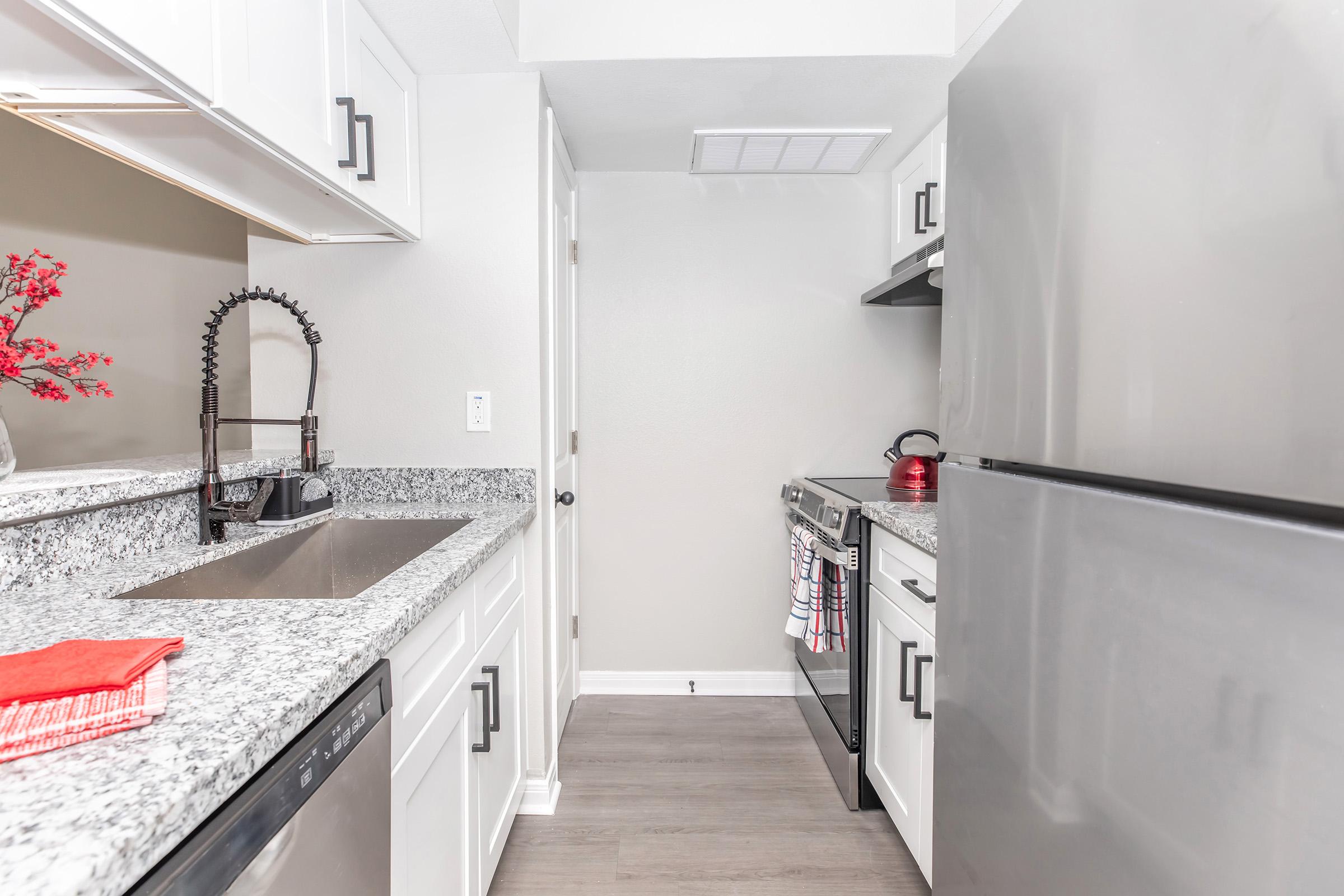 A modern kitchen featuring light gray walls, granite countertops, and stainless steel appliances. The space includes a sink with a black faucet, an oven and stovetop, a refrigerator, and a decorative red towel on the counter. A small plant adds a touch of color to the minimalist design.