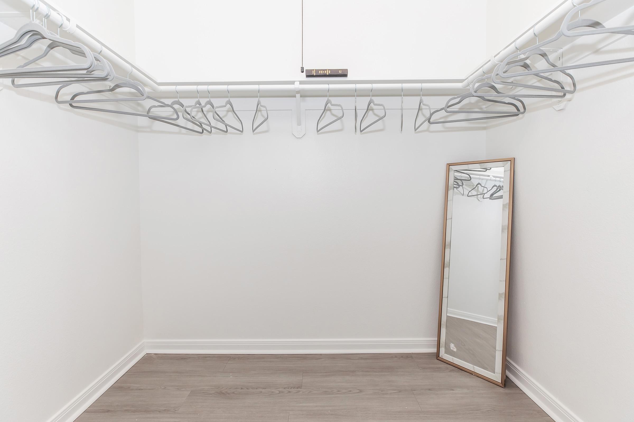 A spacious, empty closet with a white wall and light wood flooring. Several white hangers are neatly arranged on a clothing rod. A full-length mirror is leaning against the wall, reflecting the simple, uncluttered space.