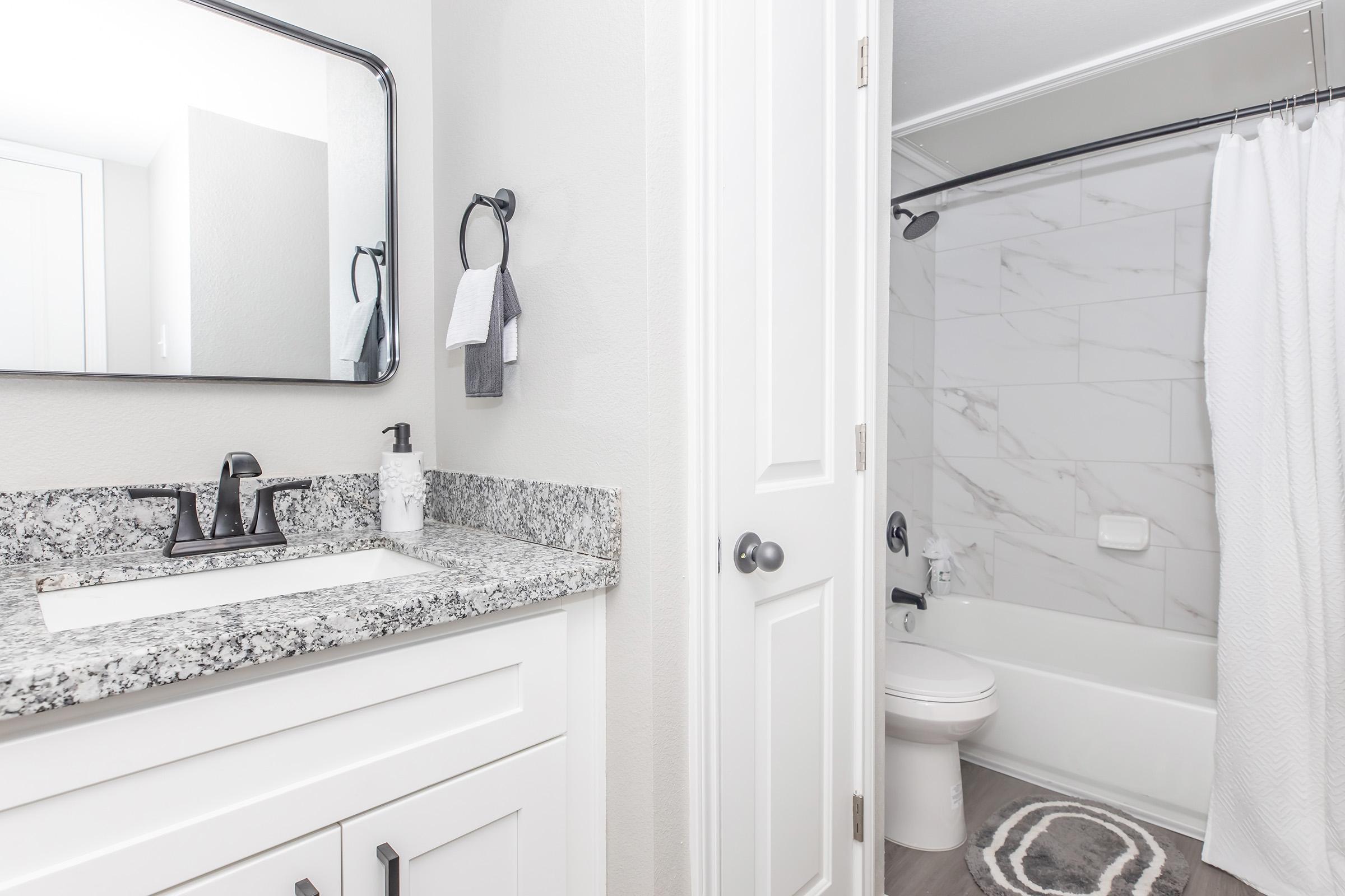 A modern bathroom featuring a white vanity with a granite countertop, a sink, and a large mirror. A towel hangs on a nearby rod, and a shower area is enclosed with a curtain. The bathroom has light-colored walls and a tiled floor, creating a clean and spacious atmosphere.