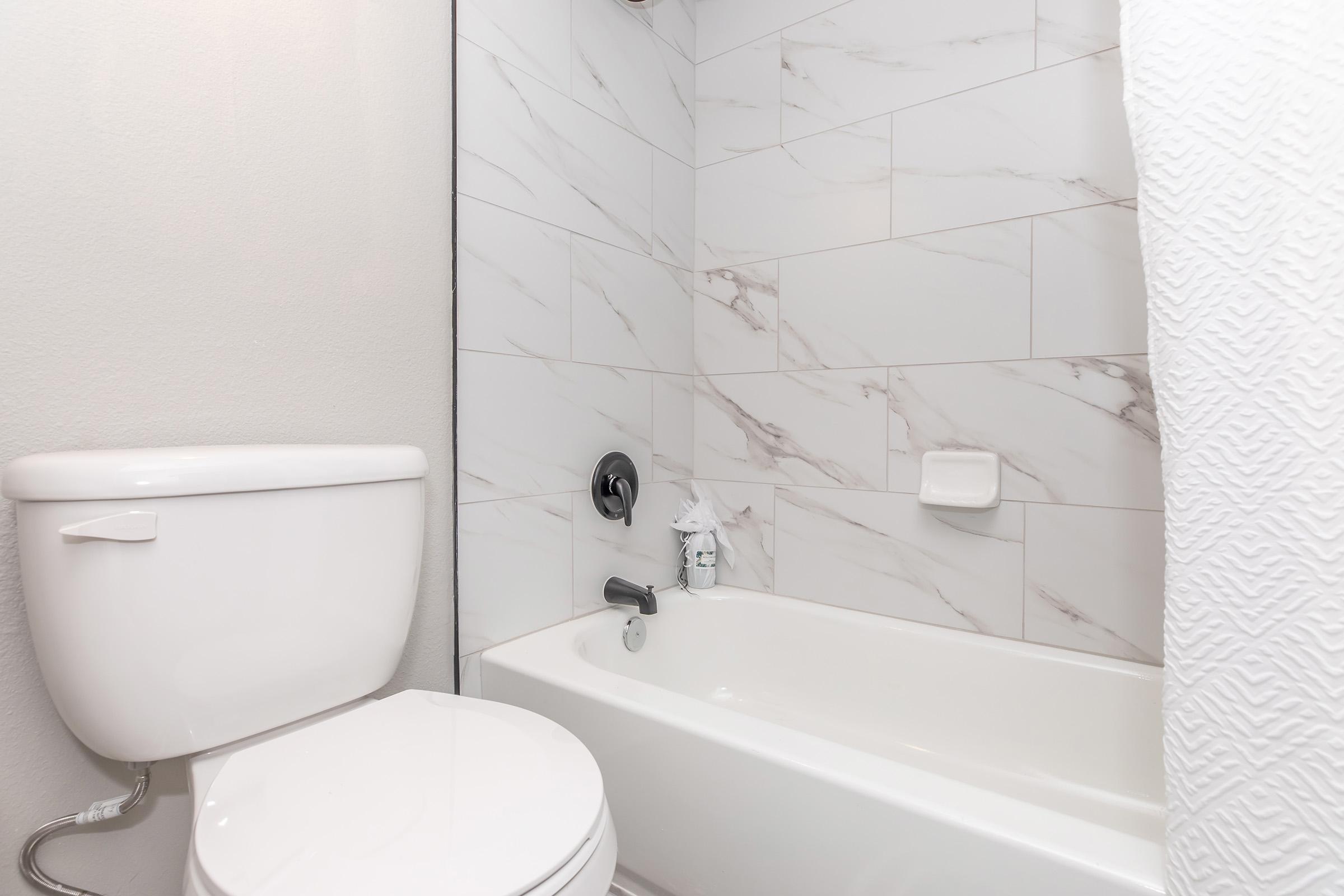 A clean, modern bathroom featuring a white toilet and a bathtub with a marble tile backdrop. The bathtub is empty, and a white shower curtain is partially visible. There is a soap holder on the wall, and a small tissue pack is placed near the faucet. The overall aesthetic is simple and contemporary.