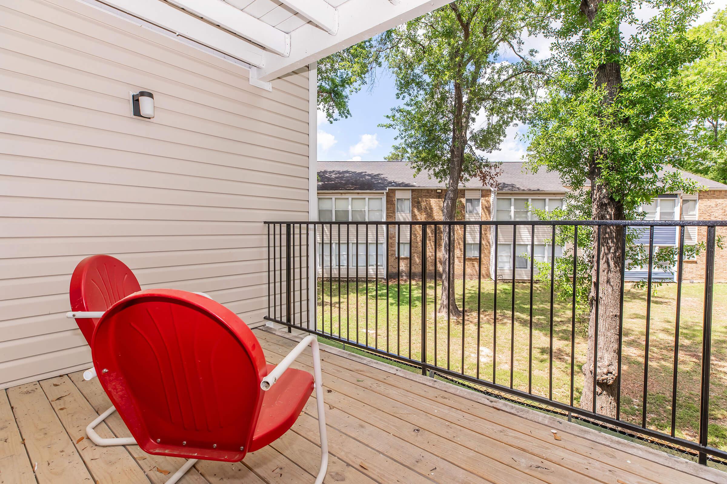 A sunny balcony with two red chairs overlooks a green courtyard with trees. The balcony features wooden flooring and white railings, with a light fixture on the wall. In the background, there's a building with multiple windows.
