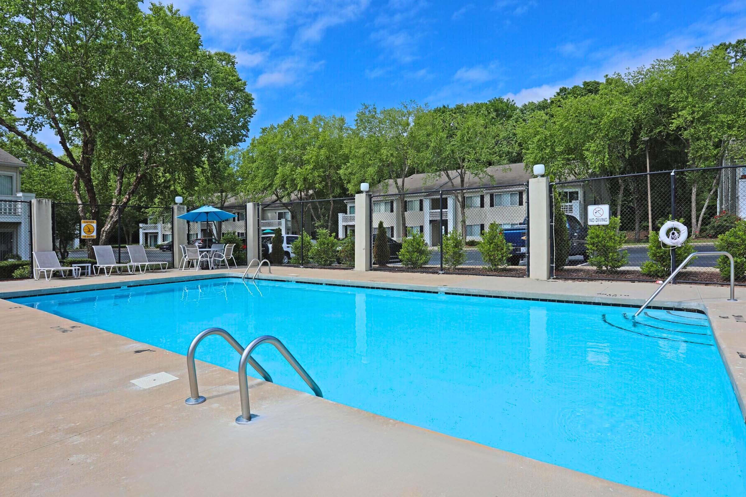 A clear, blue swimming pool surrounded by a concrete patio. There are several white lounge chairs near the pool, and a blue umbrella providing shade. In the background, a fenced area with green trees and apartment buildings is visible. The sky is bright blue with scattered clouds.