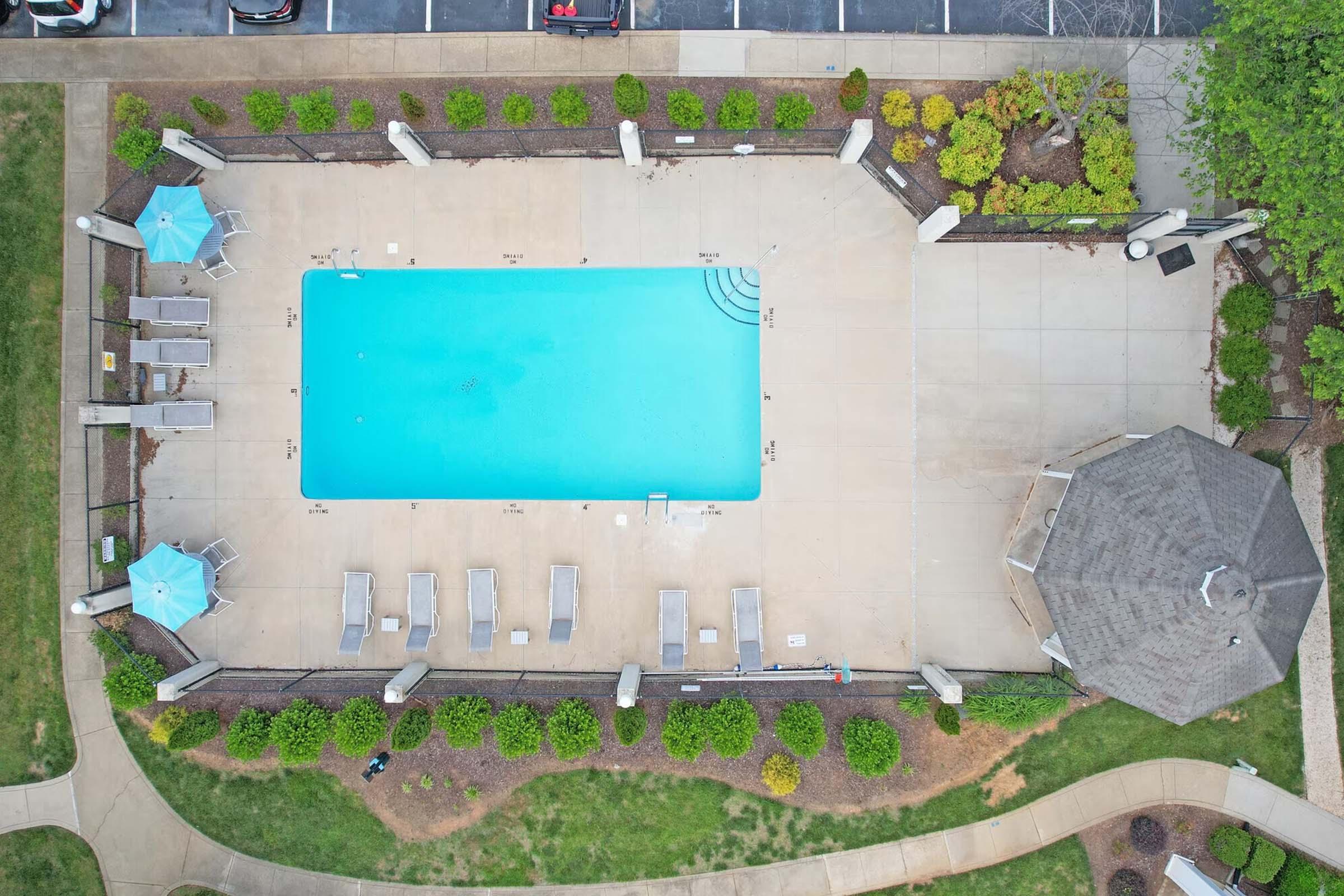 Aerial view of a swimming pool surrounded by lounge chairs and umbrellas. The pool is clear and inviting, with a gazebo nearby. Neatly landscaped areas with shrubs and greenery enhance the setting, while a parking lot is visible in the background.