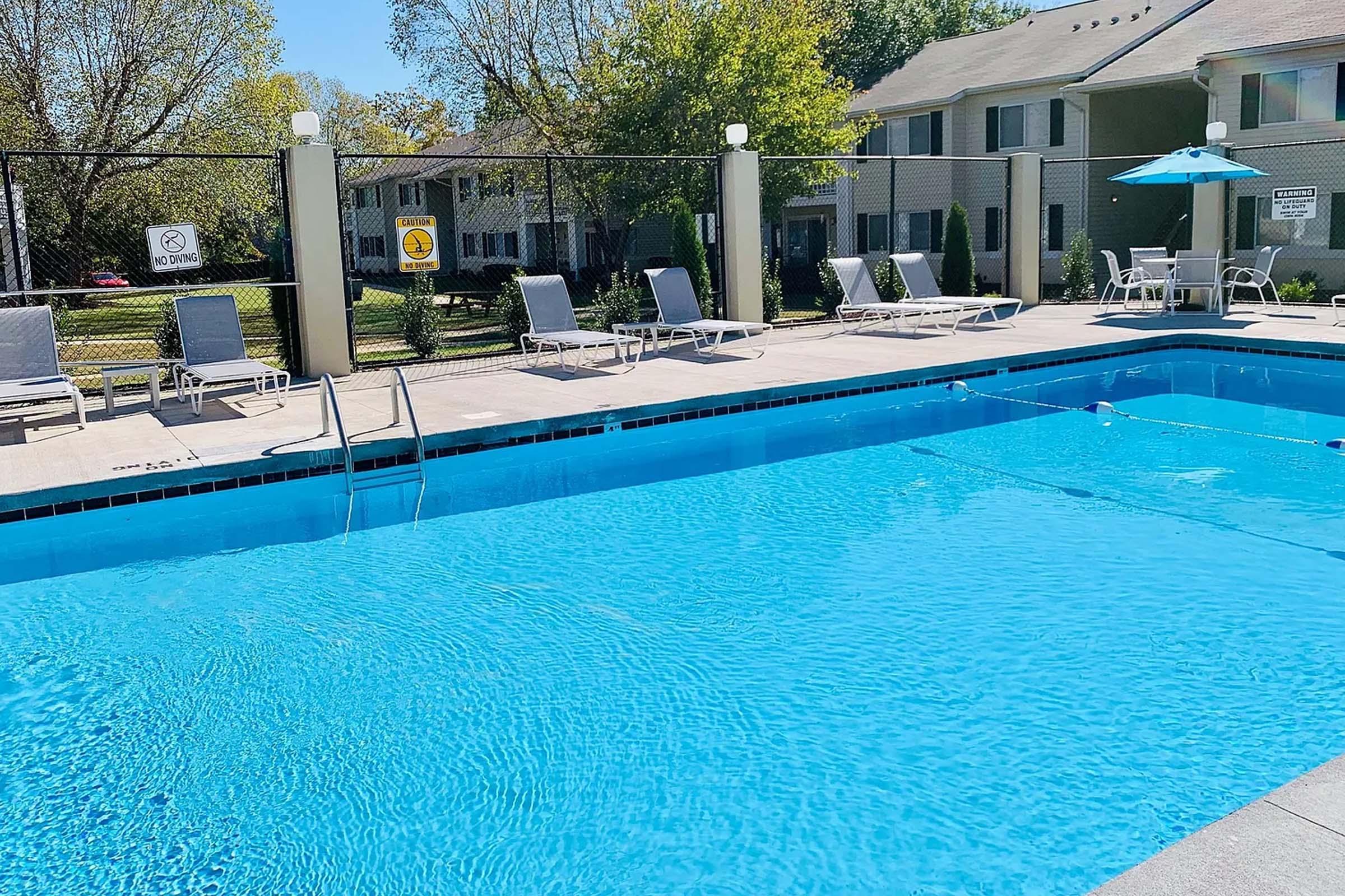 A sunny view of a swimming pool surrounded by lounge chairs. The clear blue water reflects the sunlight, and there are umbrellas for shade. Nearby, there are signs indicating pool rules. Lush trees and apartment buildings are visible in the background.